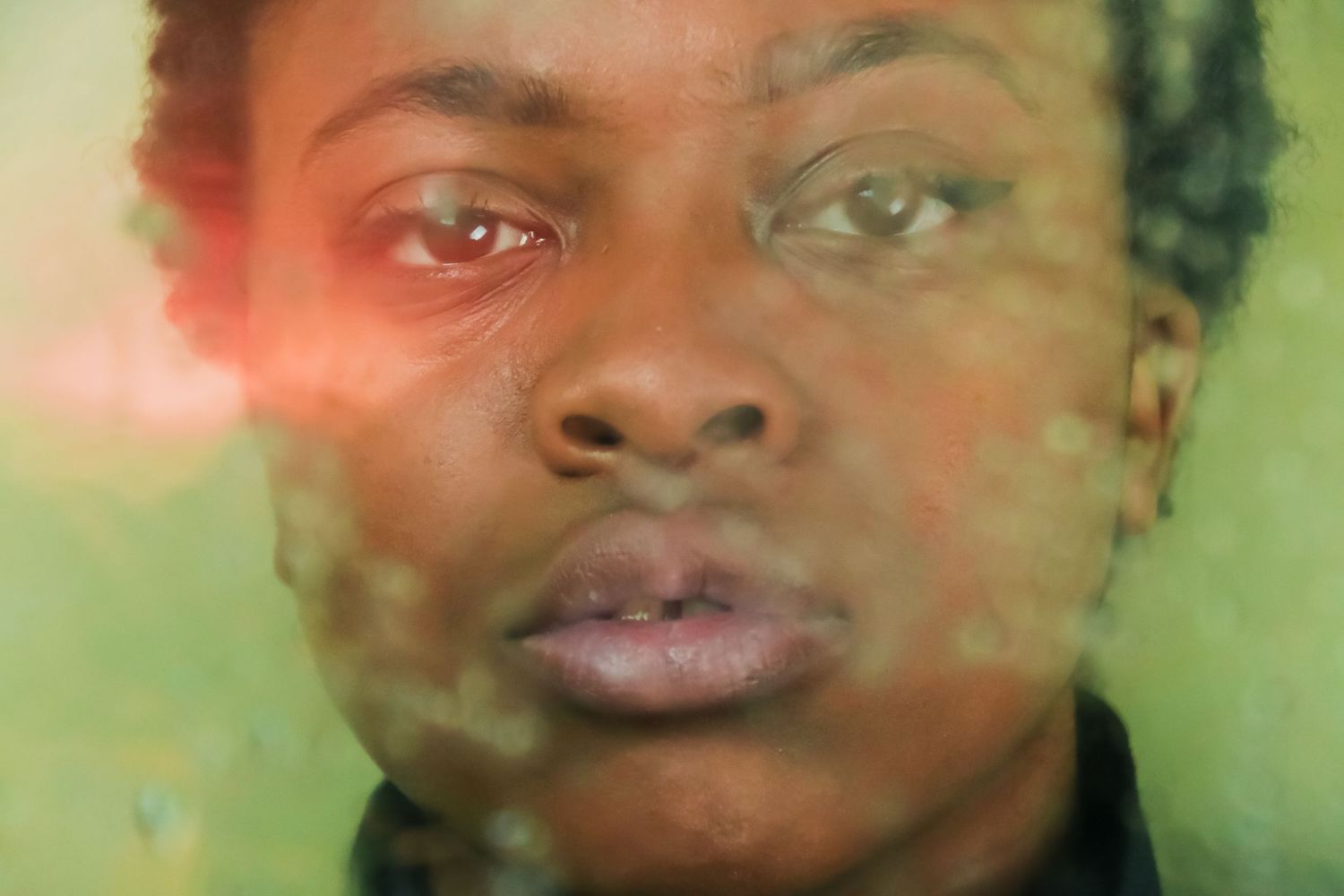 Close-up portrait of Black person's face viewed through wet green-tinted glass surface.