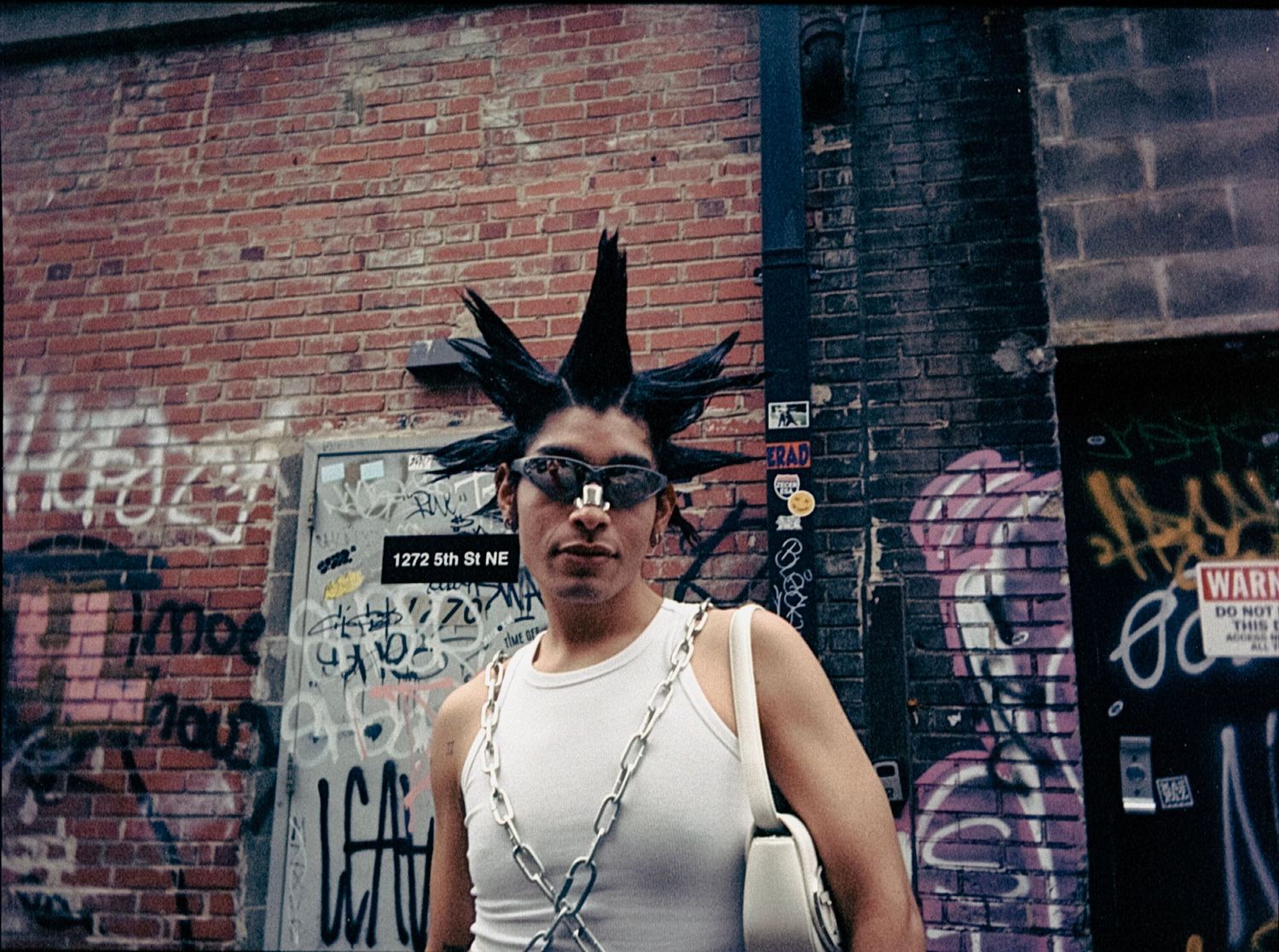 Person with dramatic black spiked mohawk and sunglasses standing before graffiti-covered brick wall.