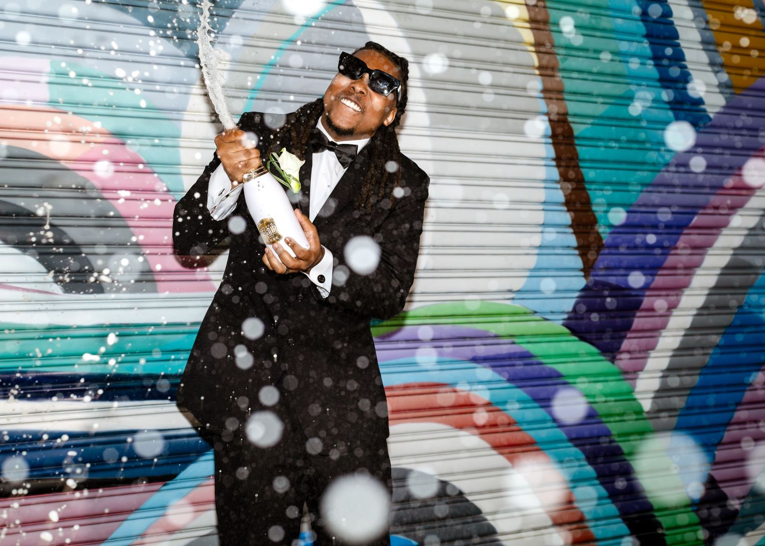 Man in a tuxedo spraying champagne in front of a colorful graffiti wall with snow falling.