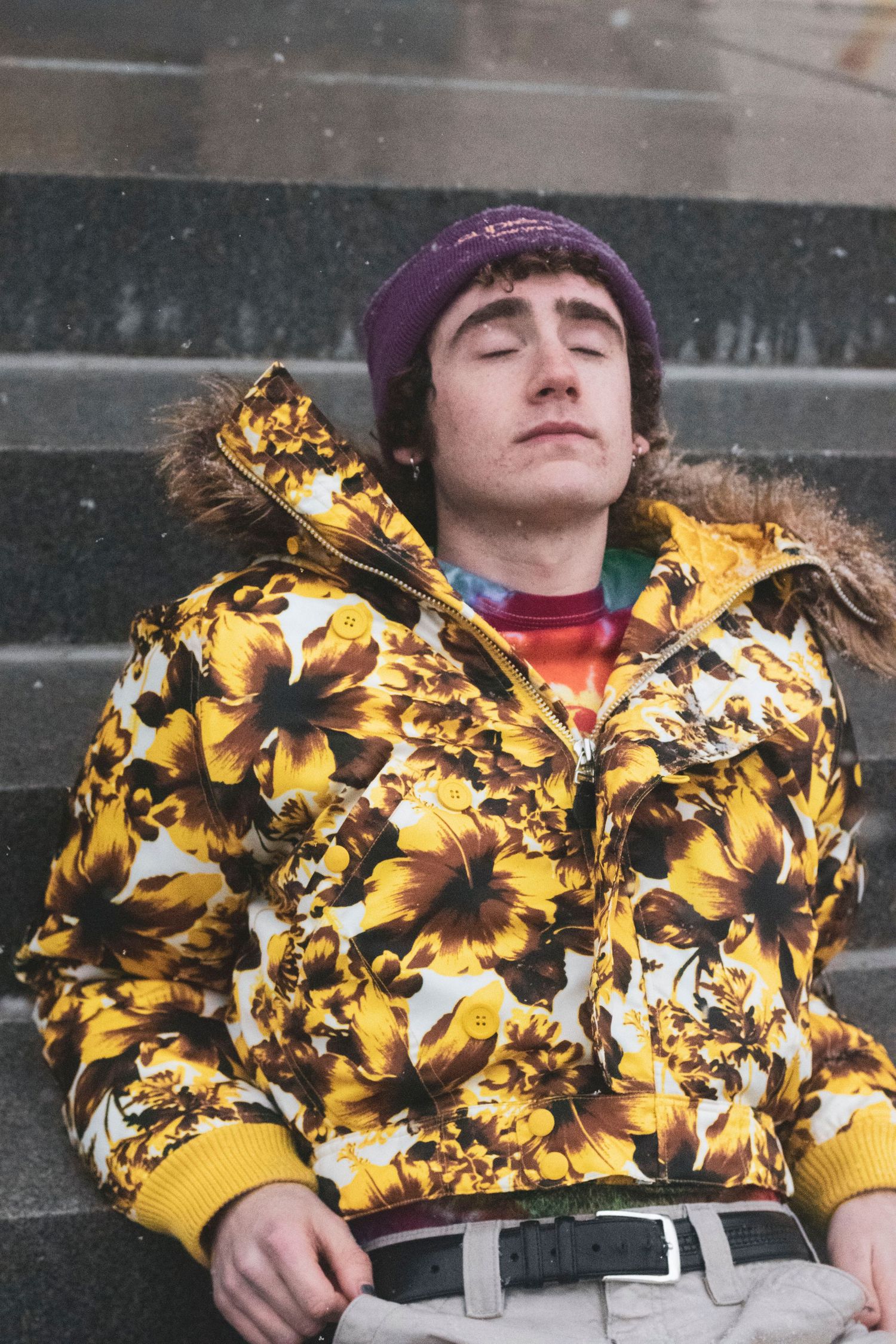 Young man in floral puffer jacket and purple Supreme beanie sits on snowy steps.