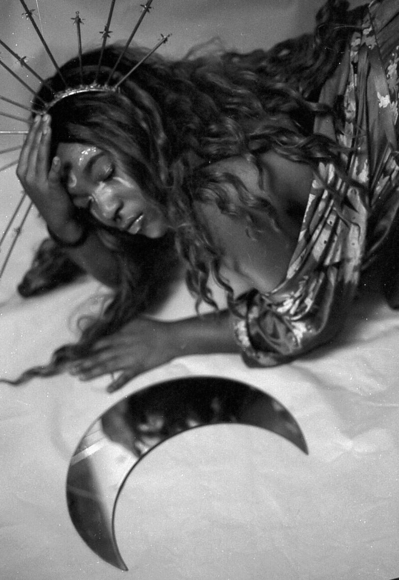 Black and white artistic photo of woman with crown and long hair posed near crescent moon prop.