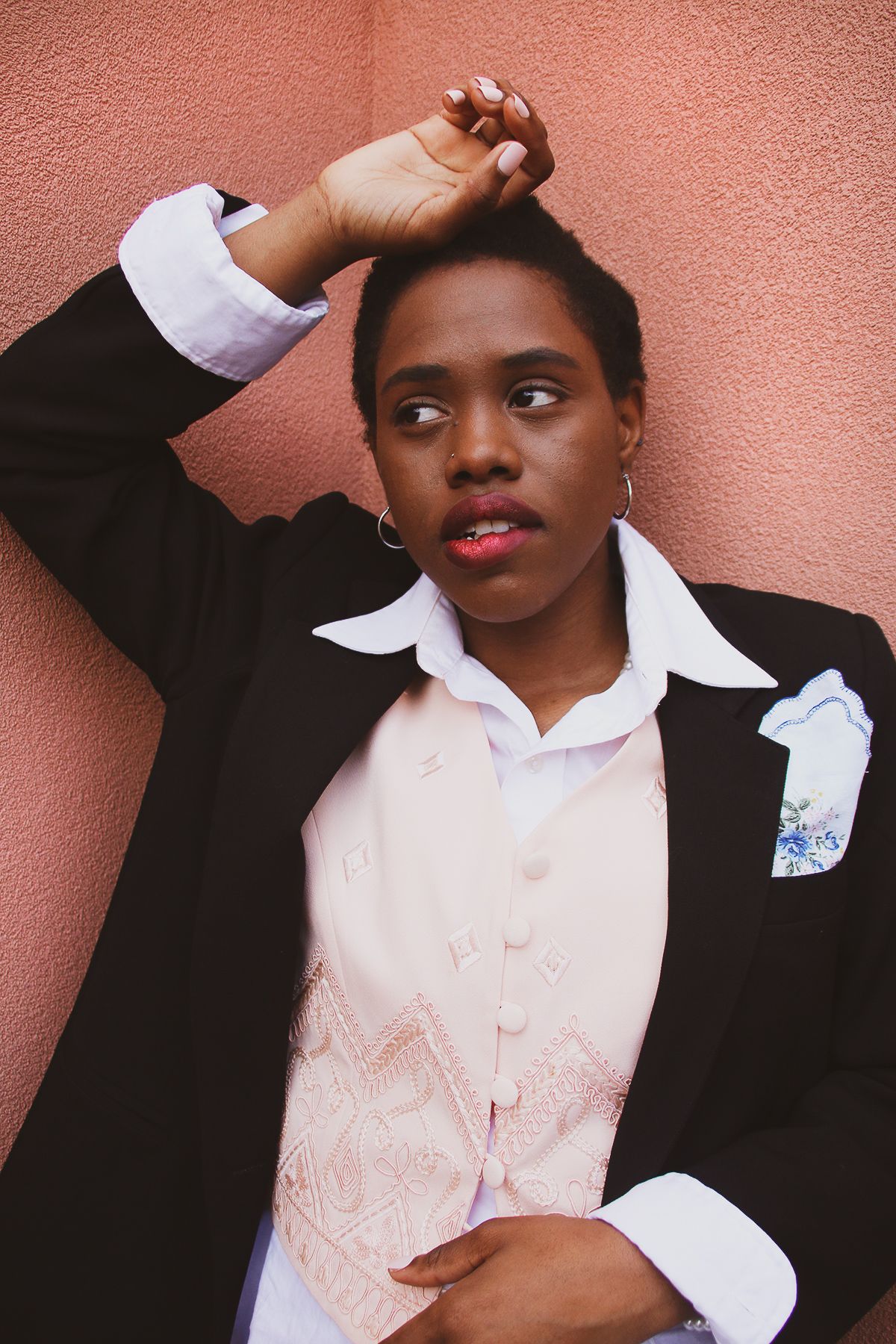 Black woman in a black blazer and pink vest posing against a pink wall.
