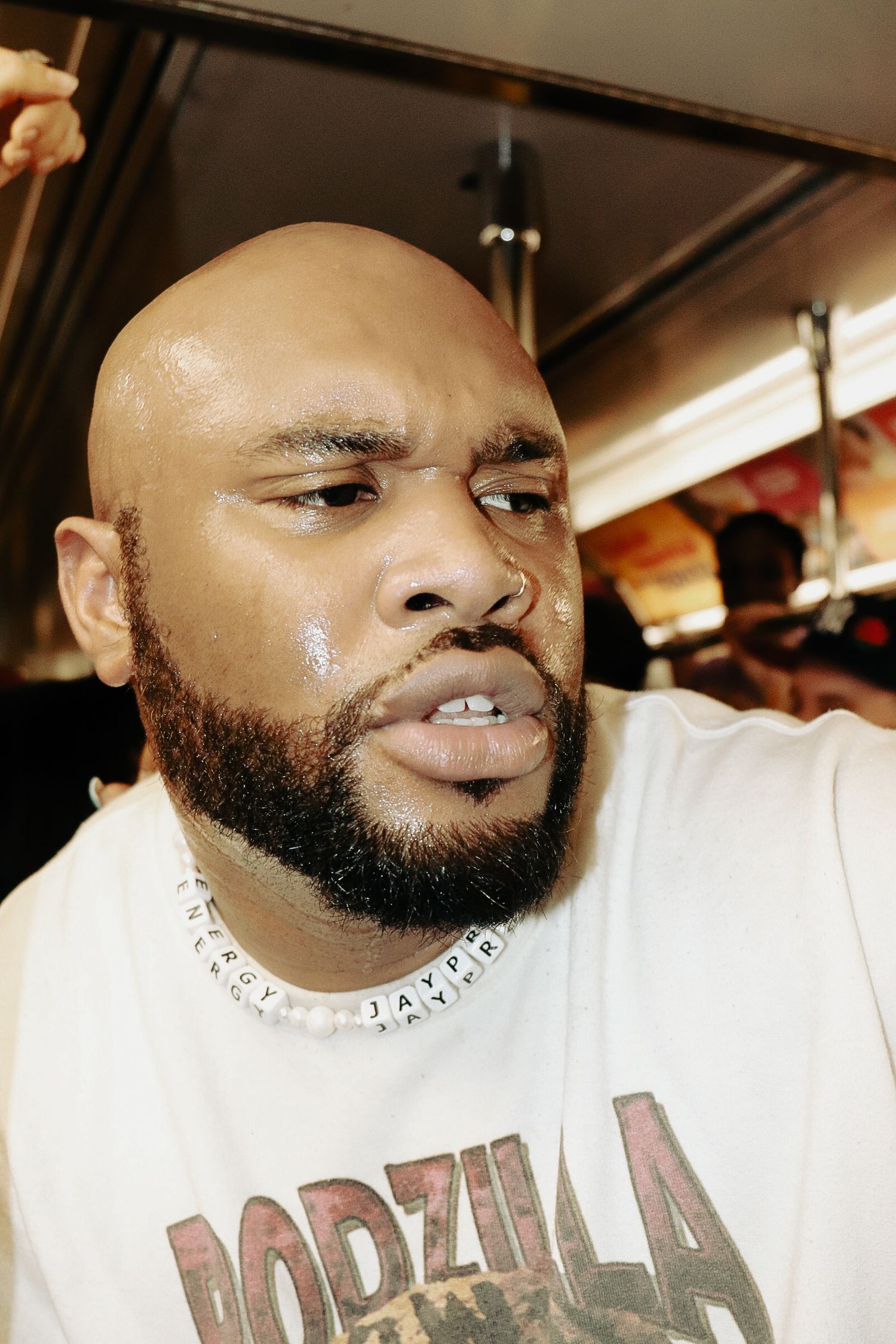 Bald man with beard wearing Godzilla t-shirt and beaded necklace sweating indoors.