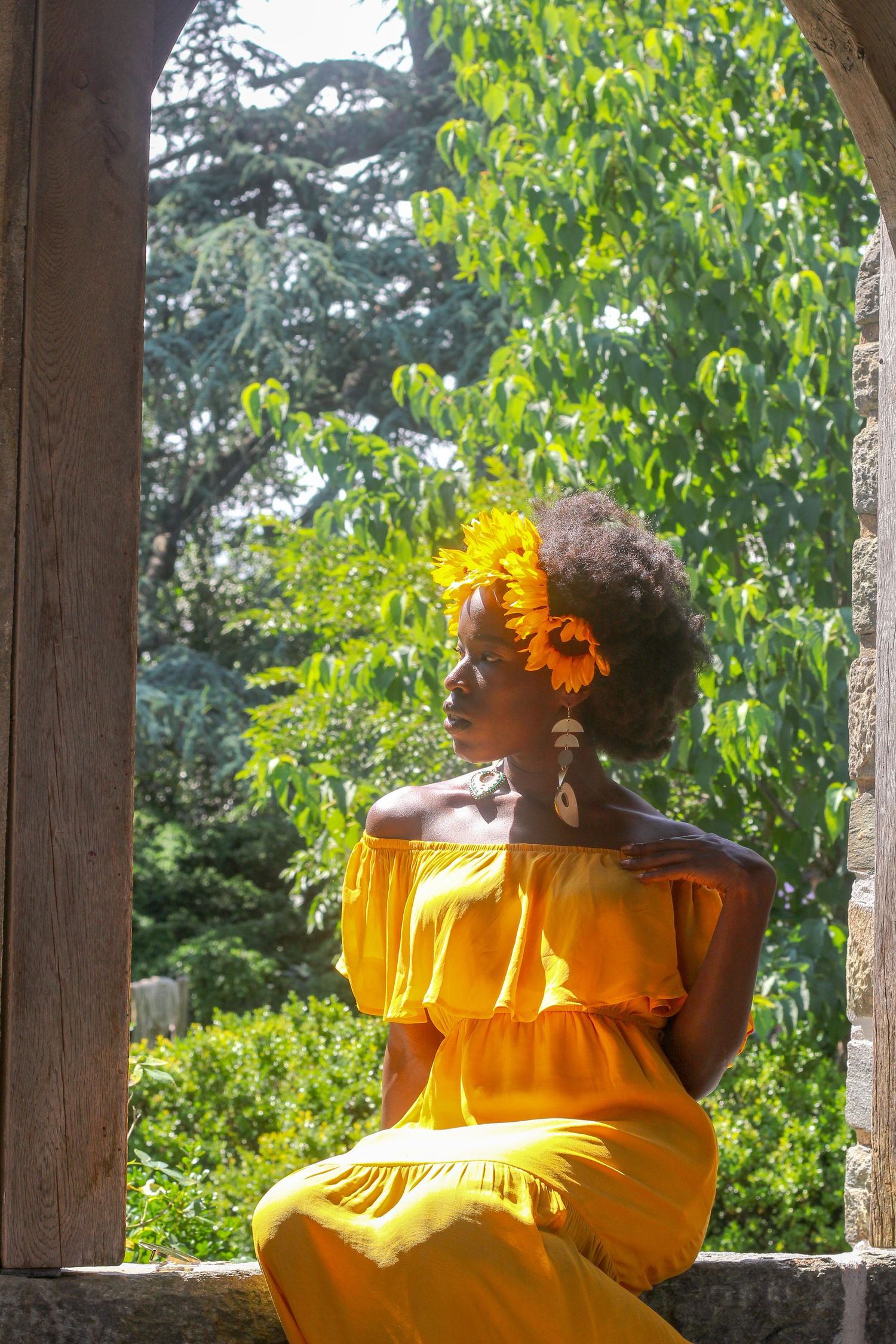 Black woman in yellow off-shoulder dress with sunflower in hair sitting in stone archway.