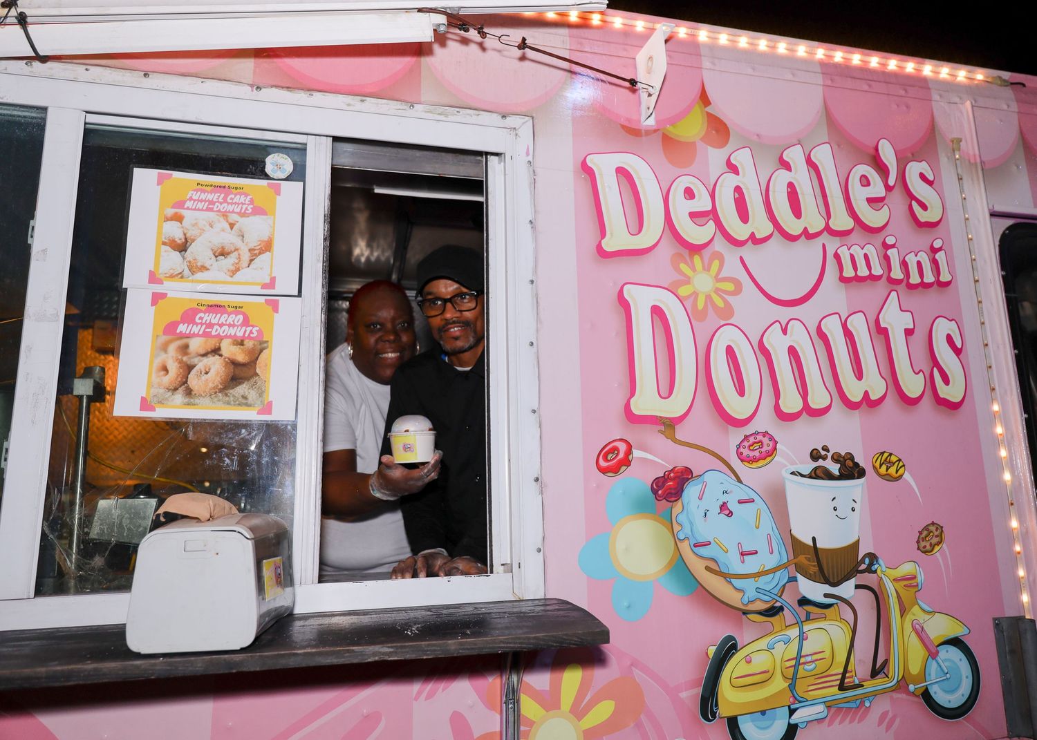 Two people smiling from window of Deddie's Mini Donuts pink food truck at night.