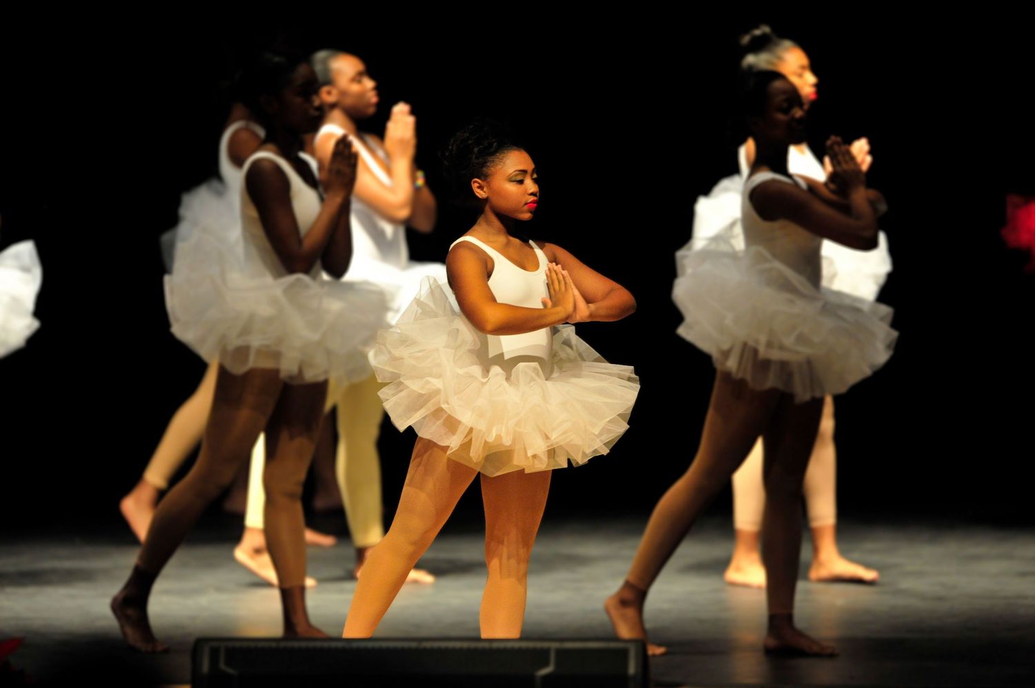 Young Black ballet dancers perform on stage in white tutus under bright stage lights.