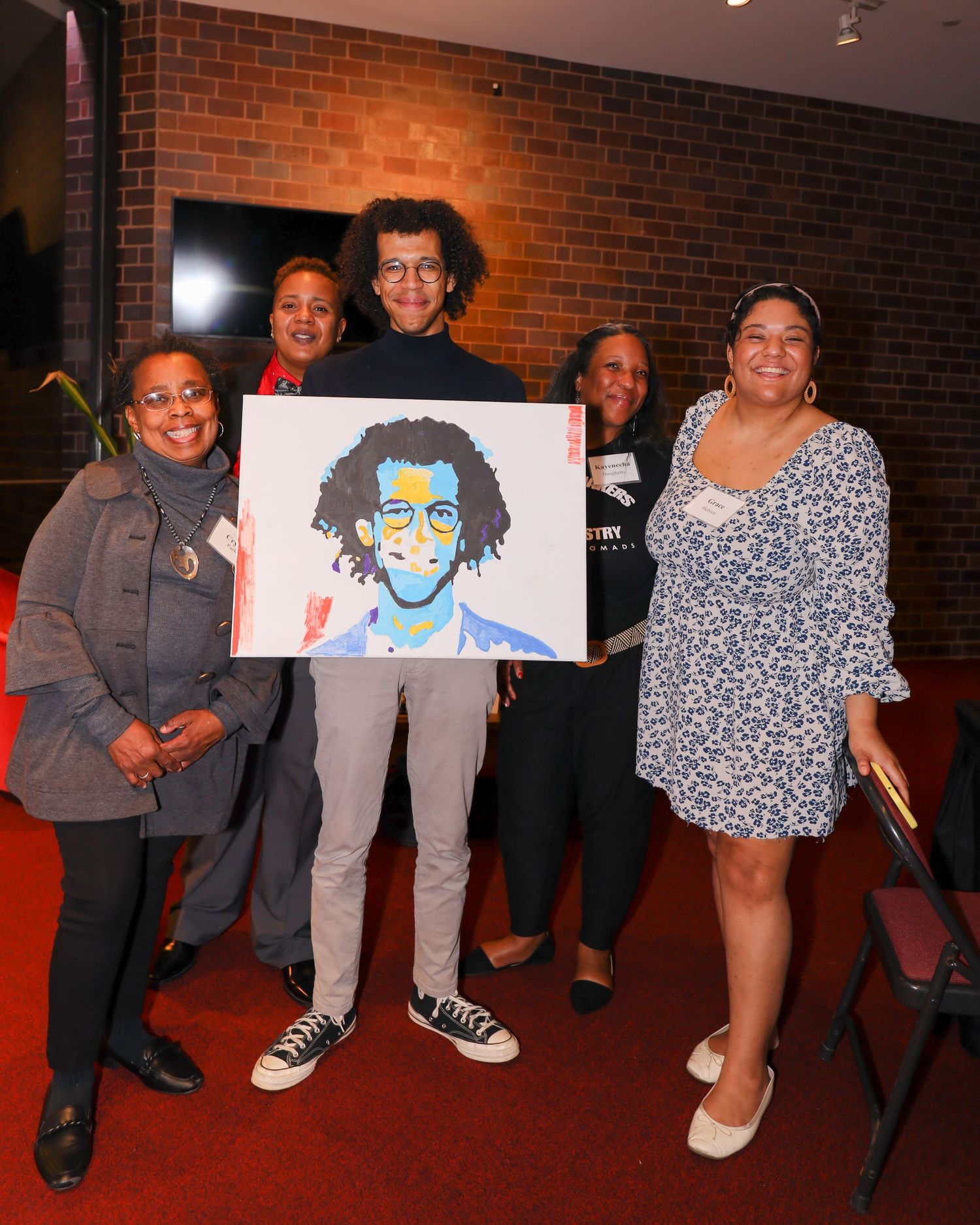 Group of five people posing with a colorful painted portrait at an indoor event.