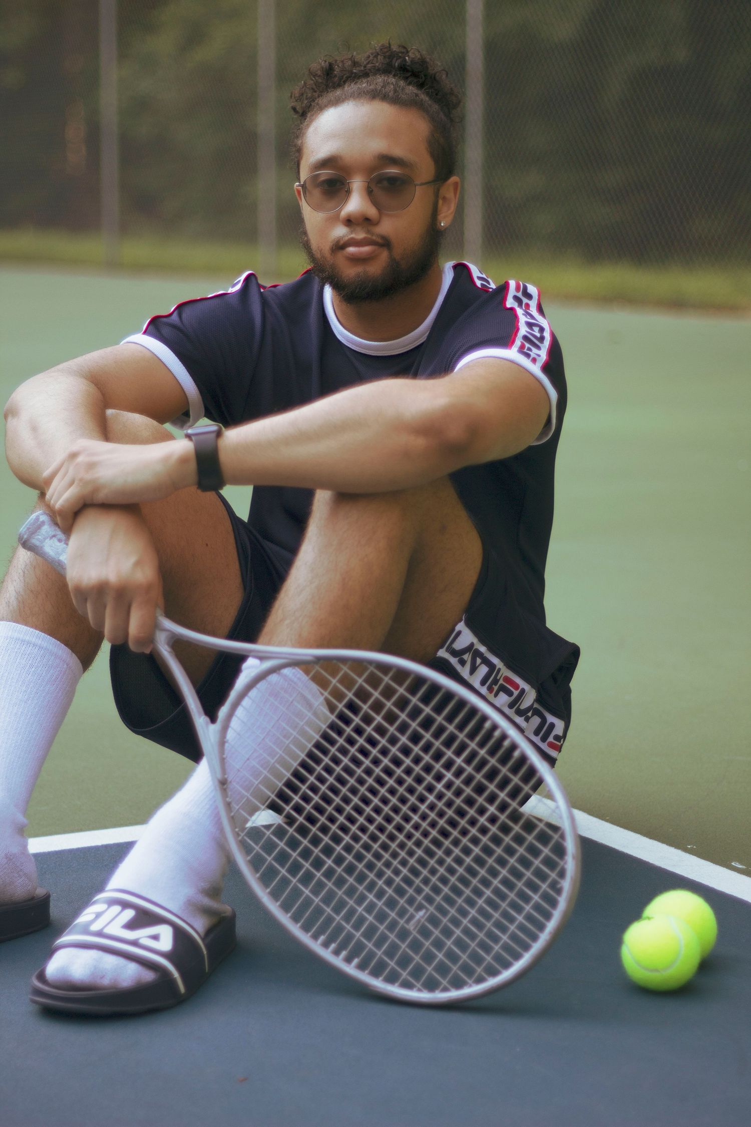 Young man with beard sitting on tennis court holding racket with tennis balls nearby.