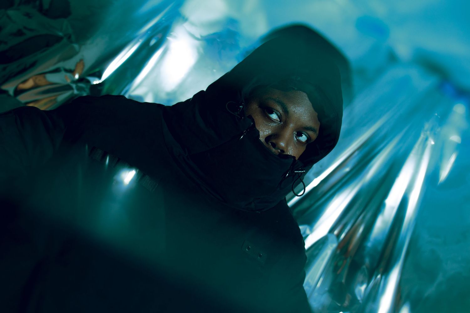 Person wearing black hood and face mask under blue-tinted lighting with reflective mylar background.