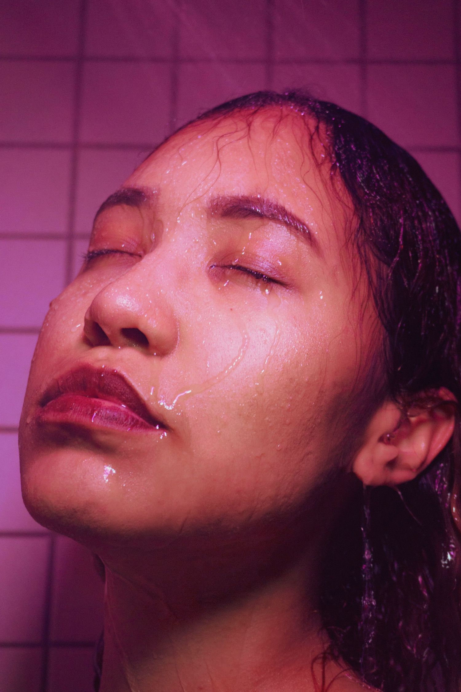 Close-up portrait of a woman with wet hair and closed eyes under pink-purple lighting with water droplets on her face.