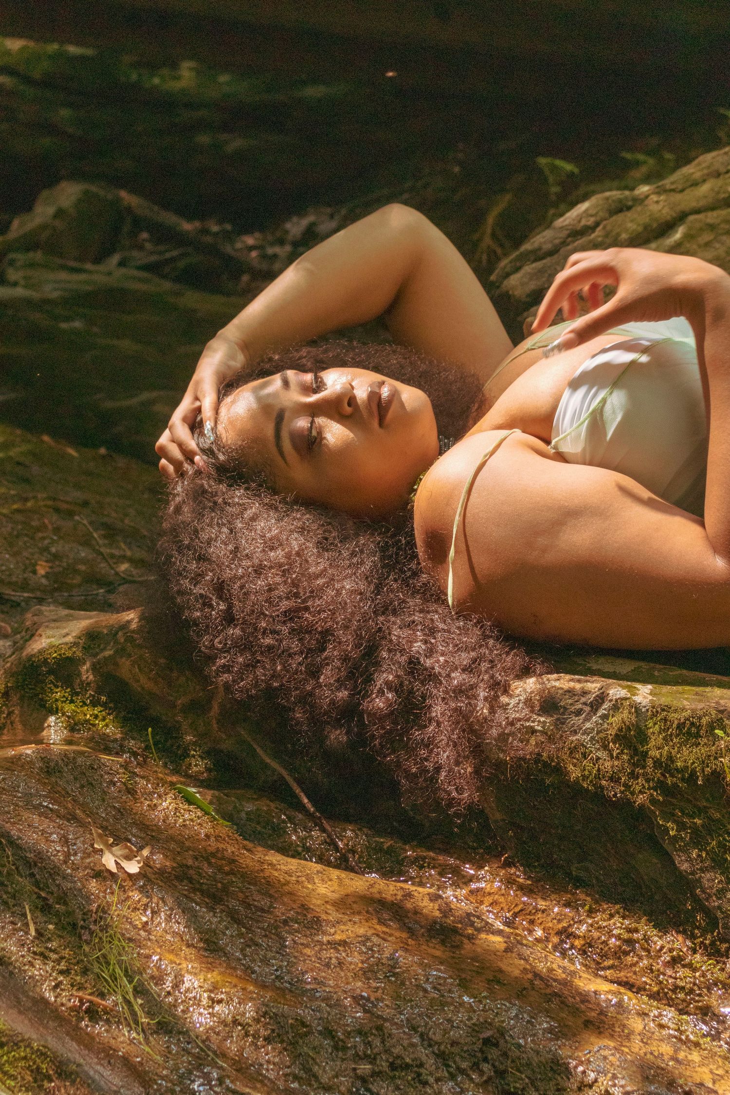 A woman with voluminous curly hair reclines on mossy rocks near a stream wearing a white bikini top.