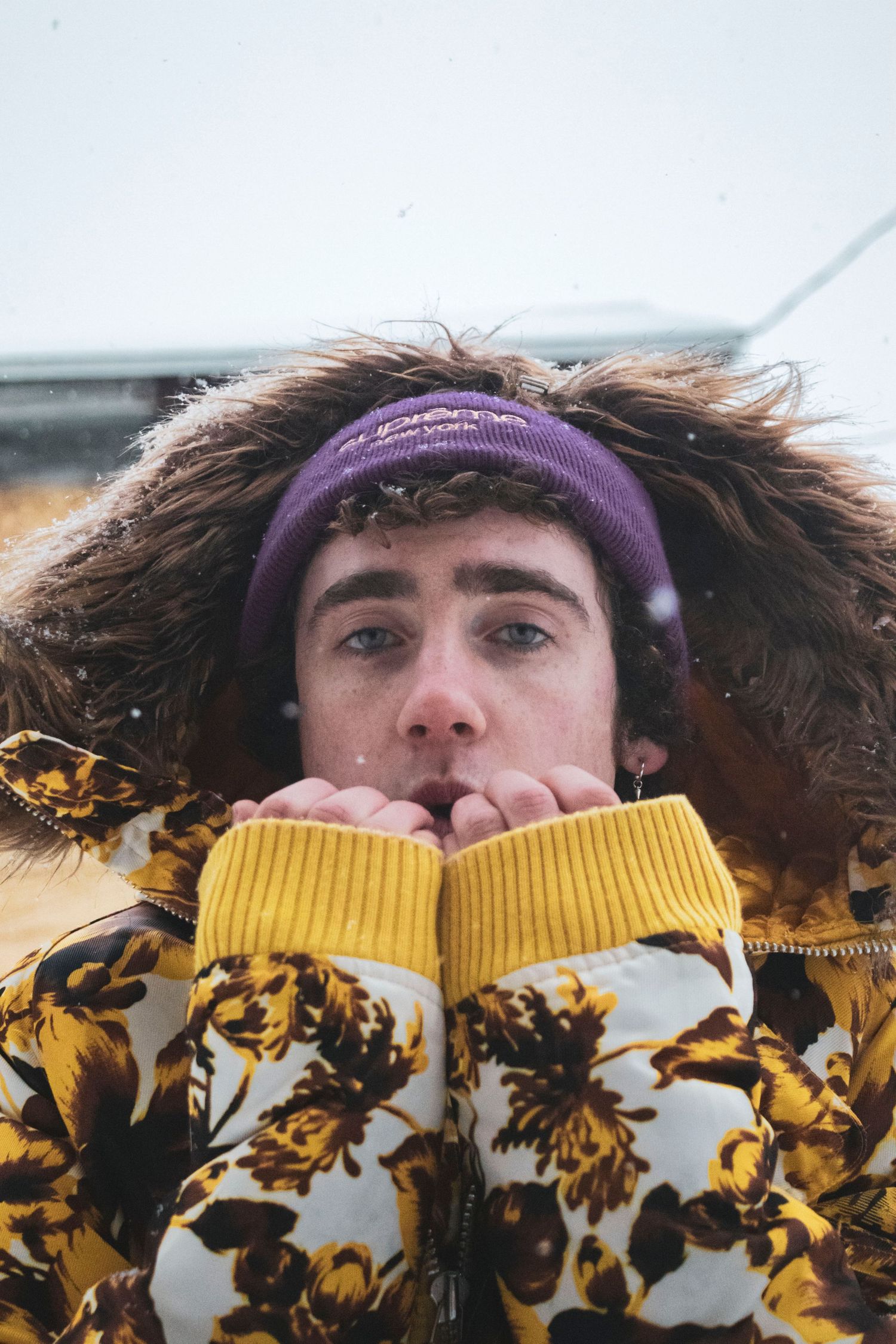 Young man in yellow floral fur-hooded jacket blows warm breath in snowy winter weather.