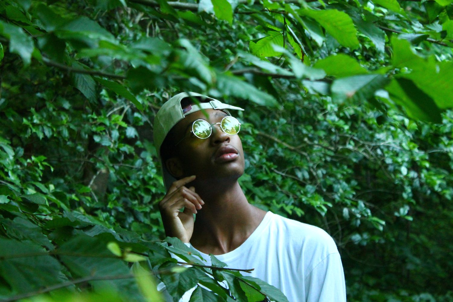 Young Black man in white tee and cap looks up through lush green tropical foliage outdoors.