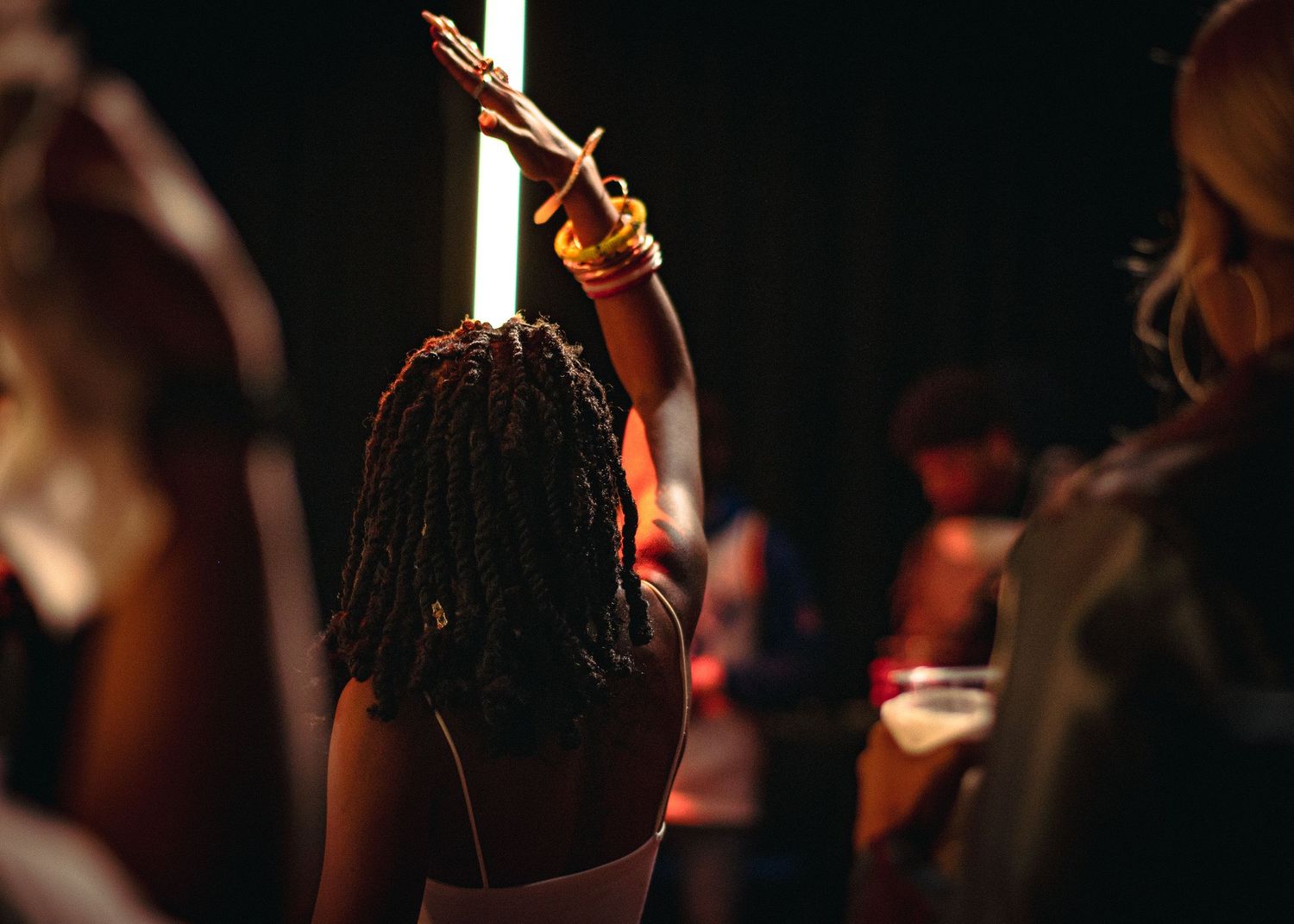 Woman with locs raising her arm holding a glowing light stick at a dark concert venue.