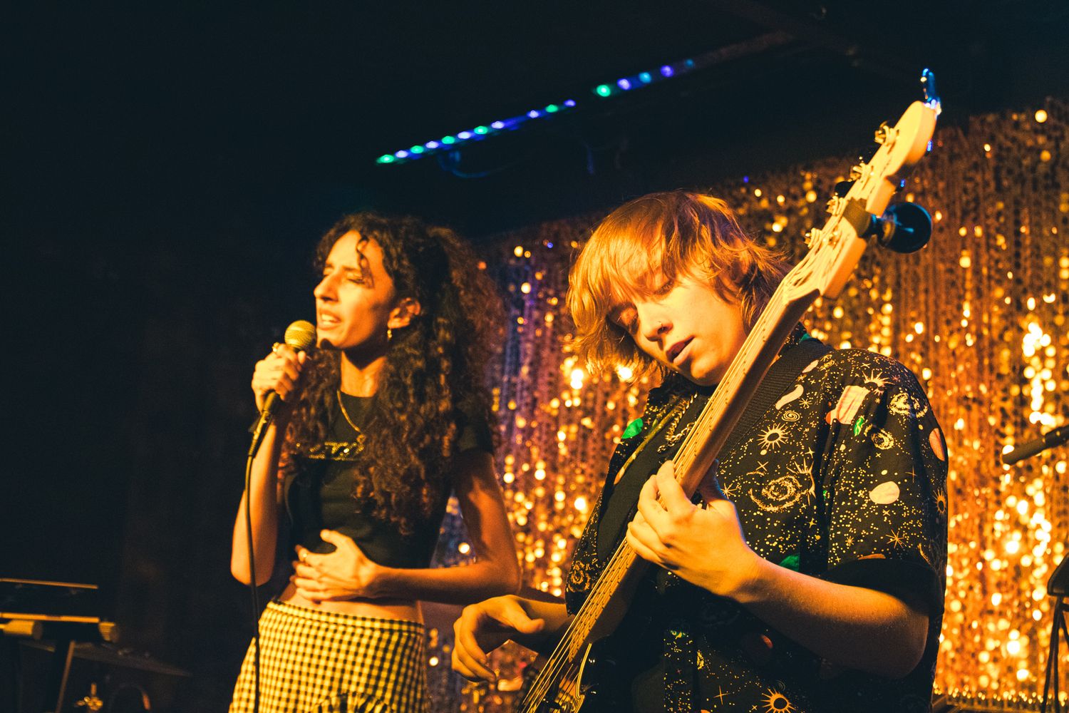 Two female musicians performing on stage with gold sequin backdrop, one singing and one playing bass guitar.