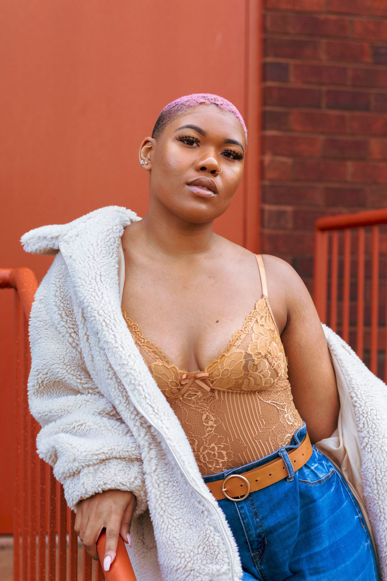 Young Black woman with purple buzzcut wearing lace bodysuit, white fleece coat, and jeans poses against red wall.