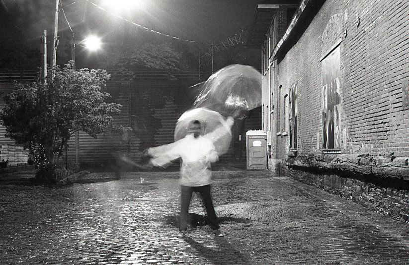 Black and white photo of person walking in rain holding umbrella on wet cobblestone alley at night.