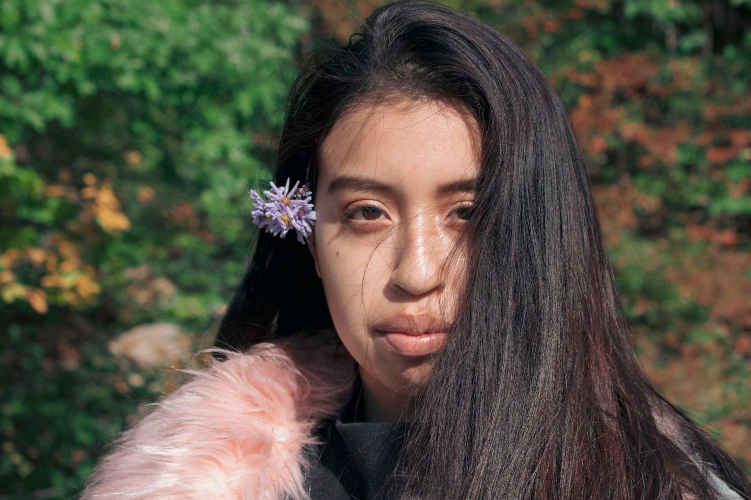 Young woman with long dark hair and a purple flower poses outdoors with a pink fur coat.