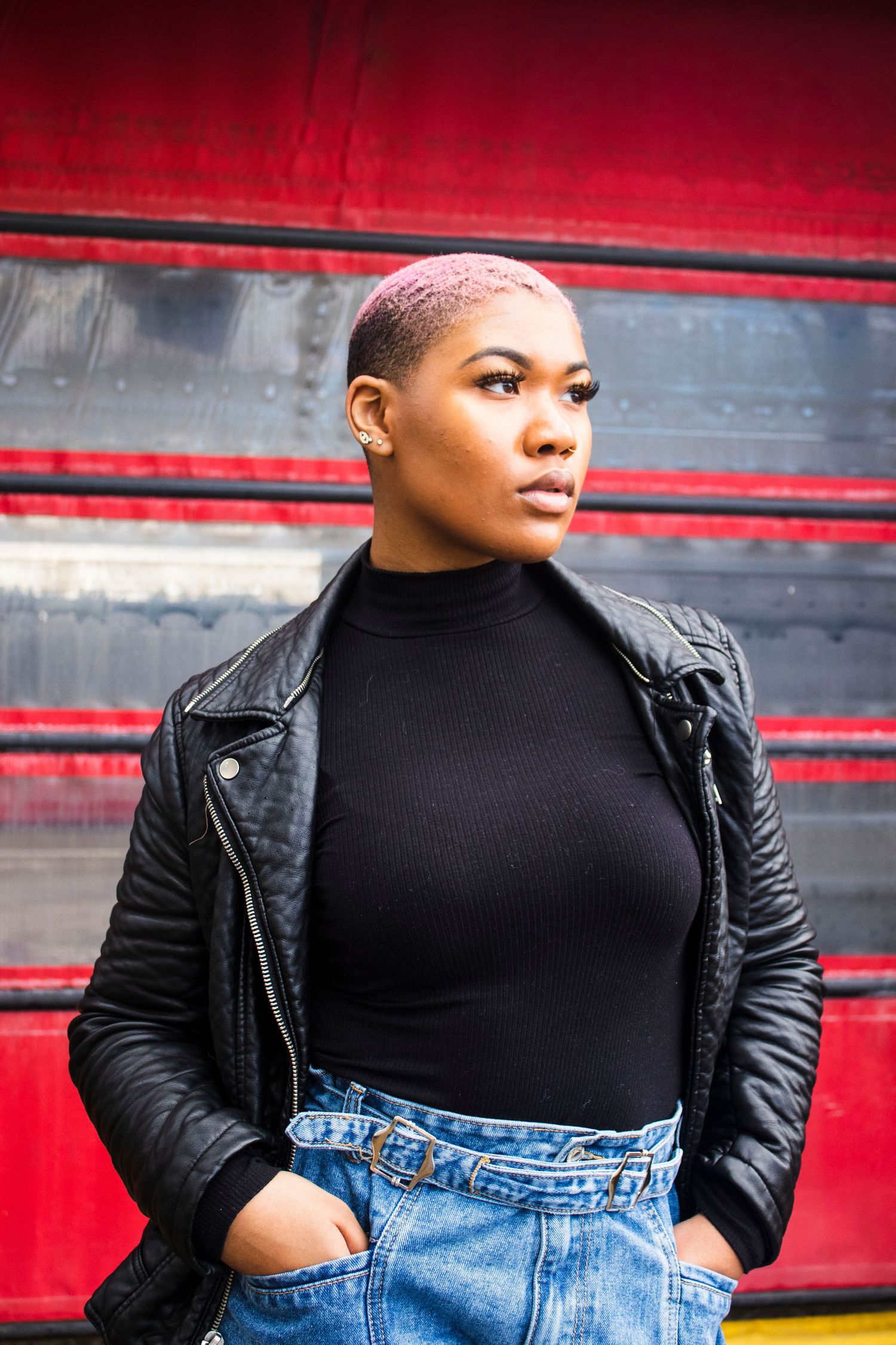 Black woman with pink buzzcut in black turtleneck and leather jacket posing against red striped background.