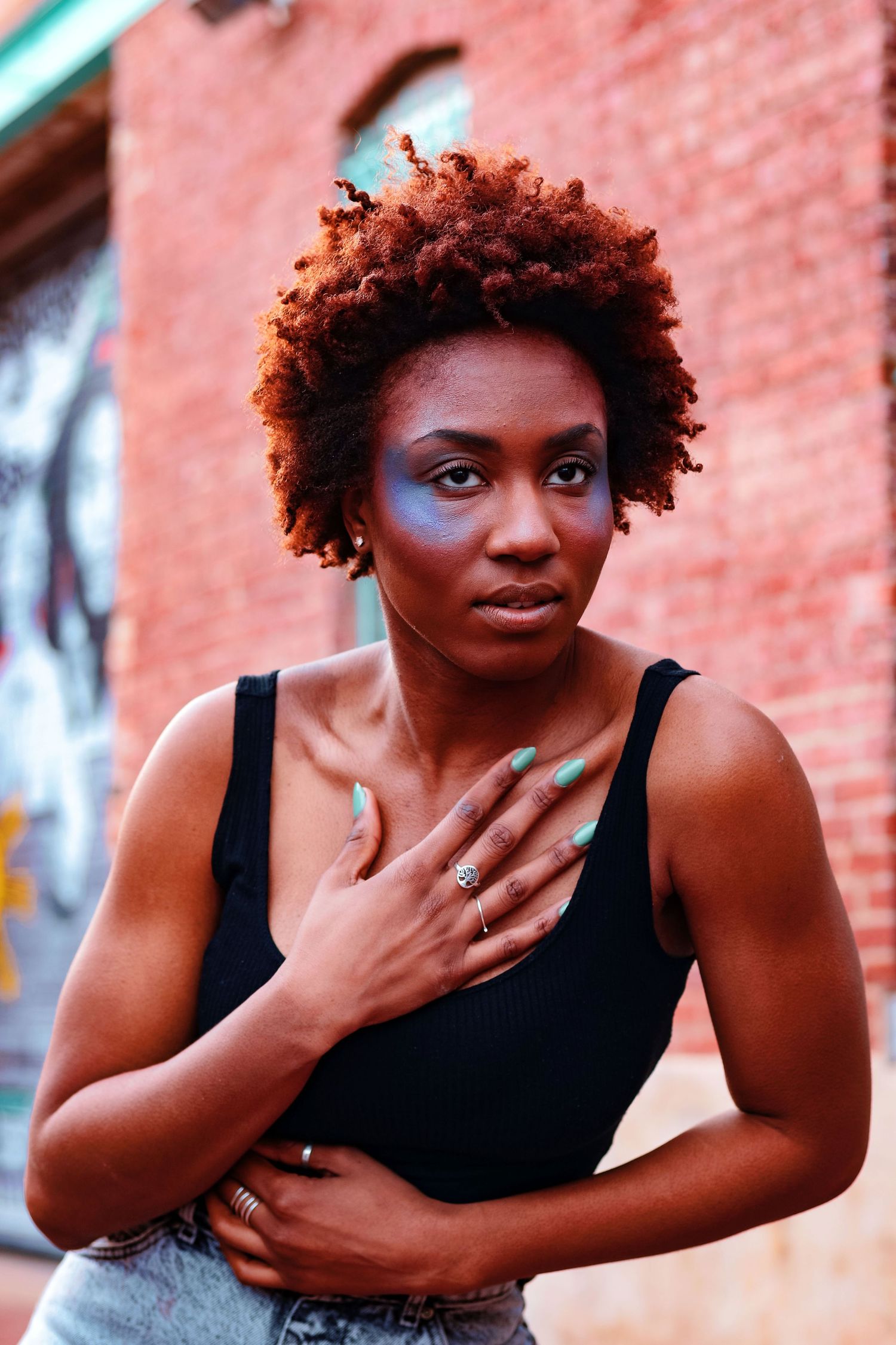 A Black woman with auburn natural hair and blue eye makeup places her hand on her chest near a brick wall.