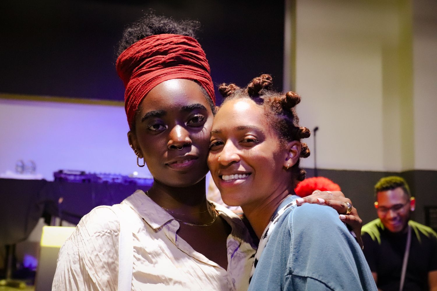Two young women smiling together at an indoor music event with warm ambient lighting.