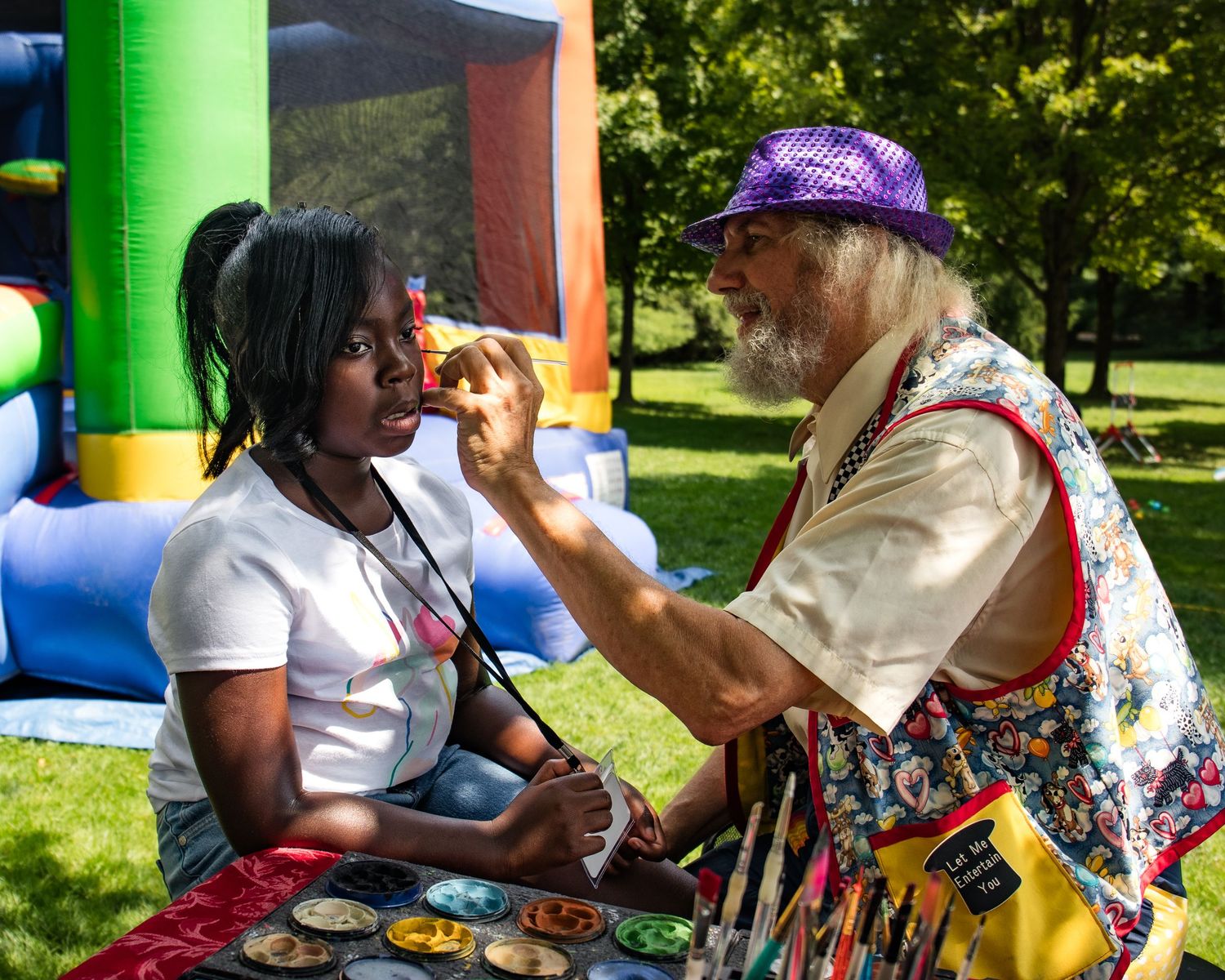 A clown artist applies colorful face paint to a young woman at an outdoor festival event.