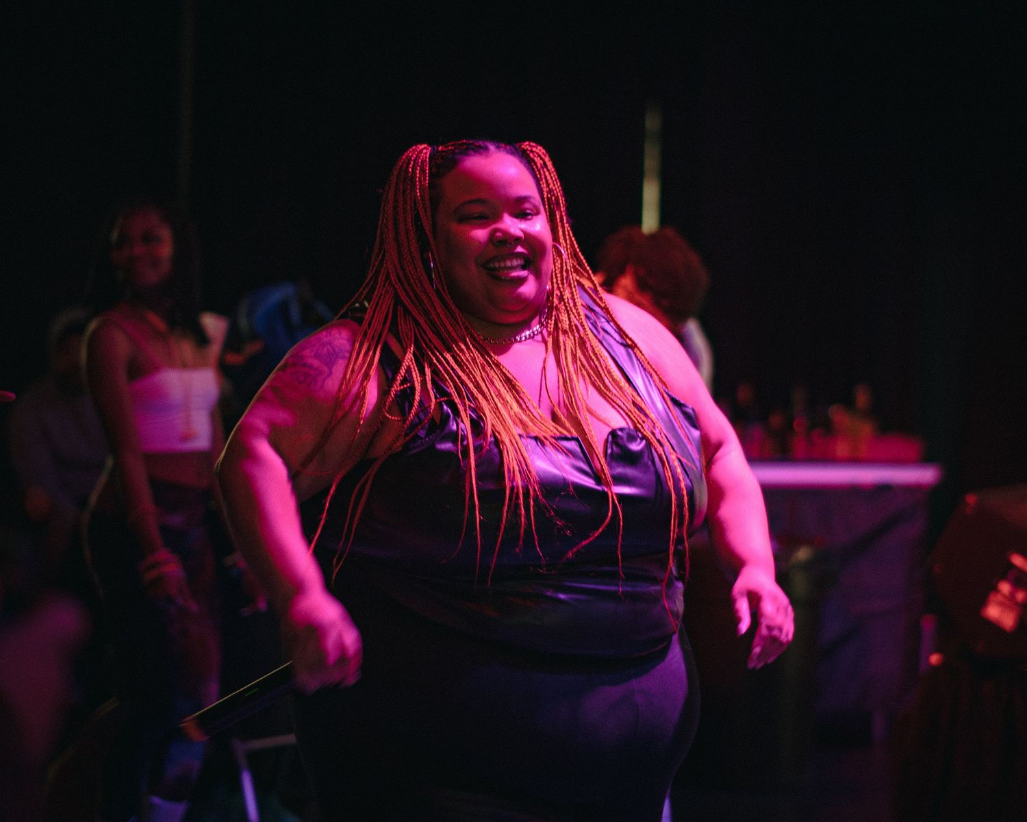 Woman with orange box braids dancing and smiling under pink stage lights at a live event.
