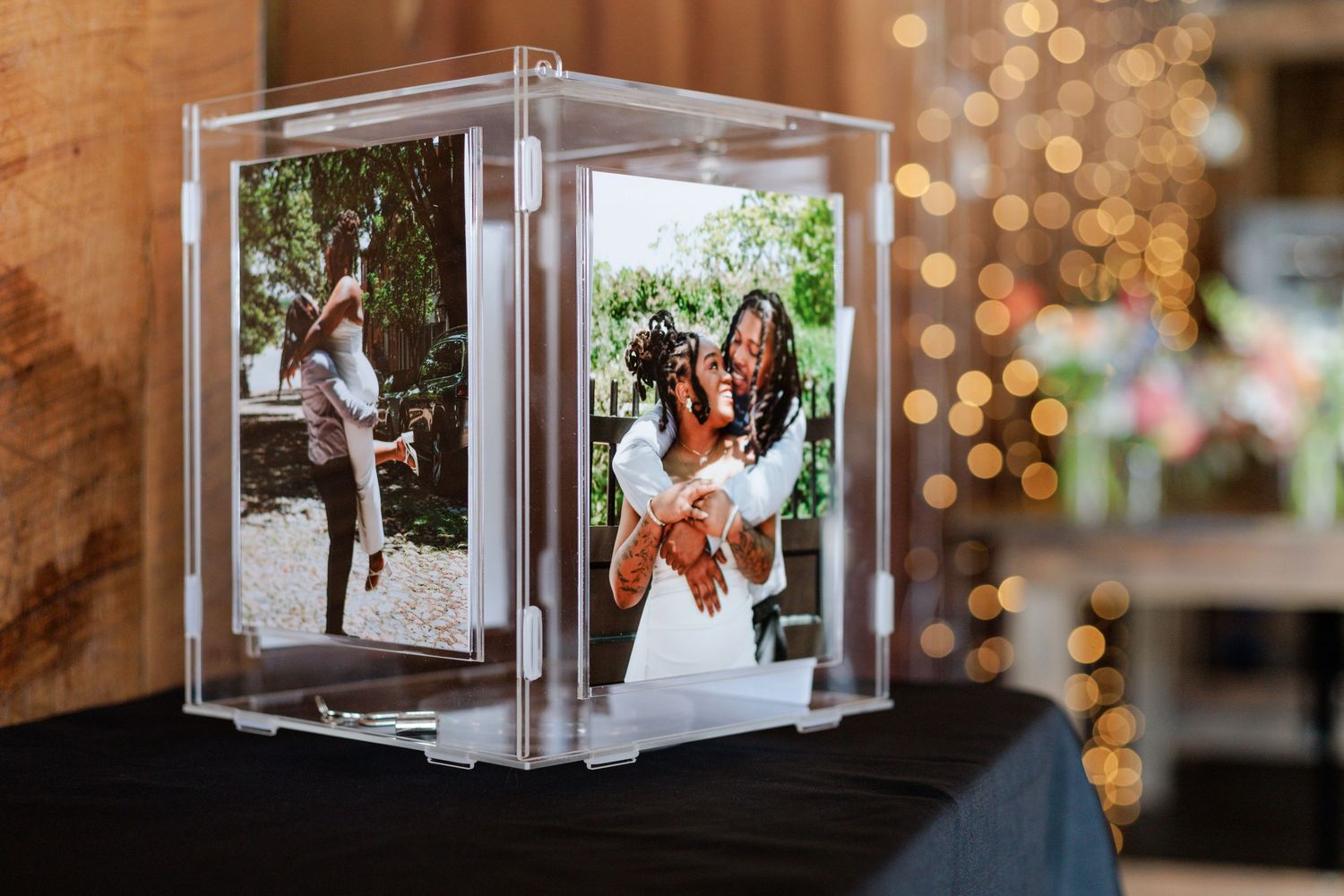 Acrylic photo display box showcasing wedding photos of two women embracing, set on a table with bokeh lights in background.
