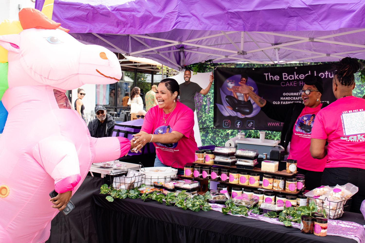 Woman in pink shirt serves customers at The Baked Lotus booth beside a large inflatable pink unicorn.