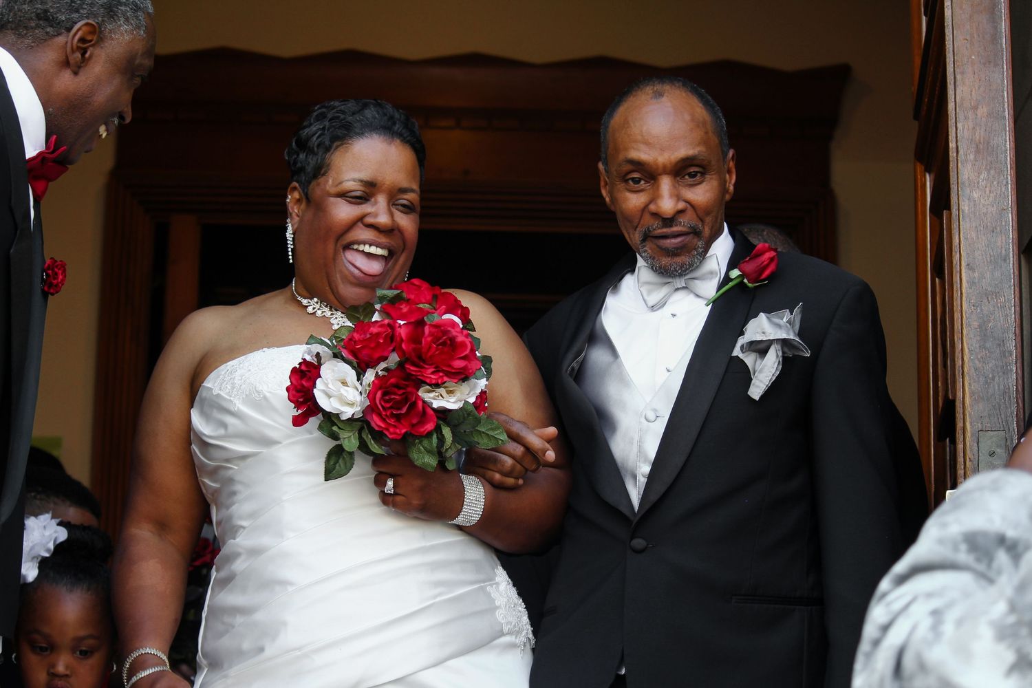 Joyful bride and groom smiling at wedding ceremony, bride holding red roses bouquet in white dress.