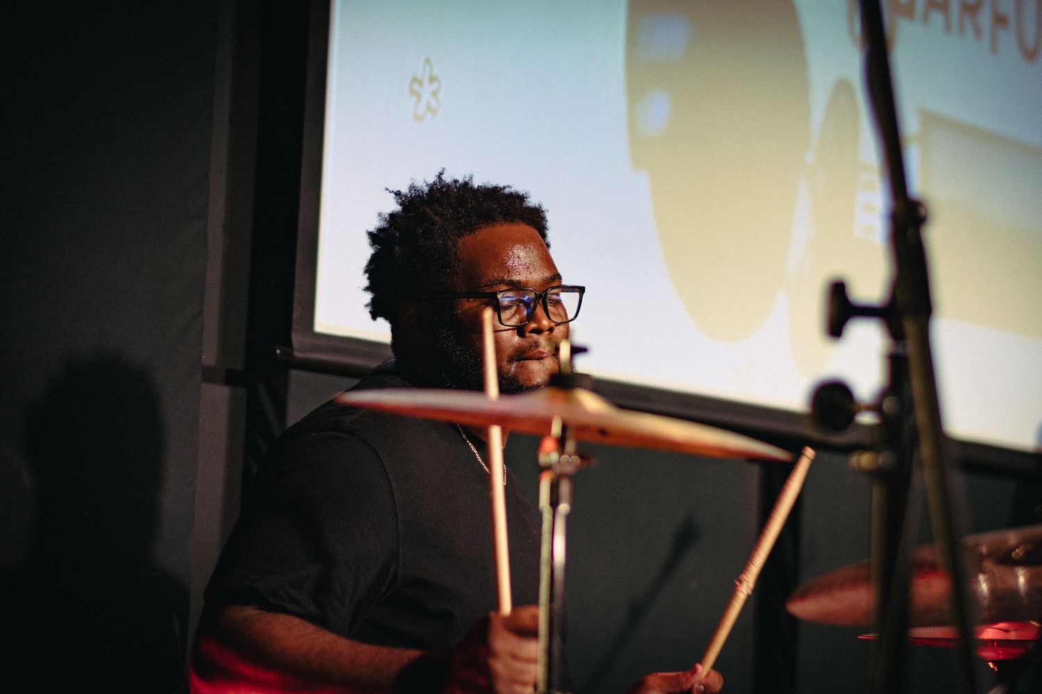 A drummer wearing glasses plays cymbals on stage with a projection screen in the background.