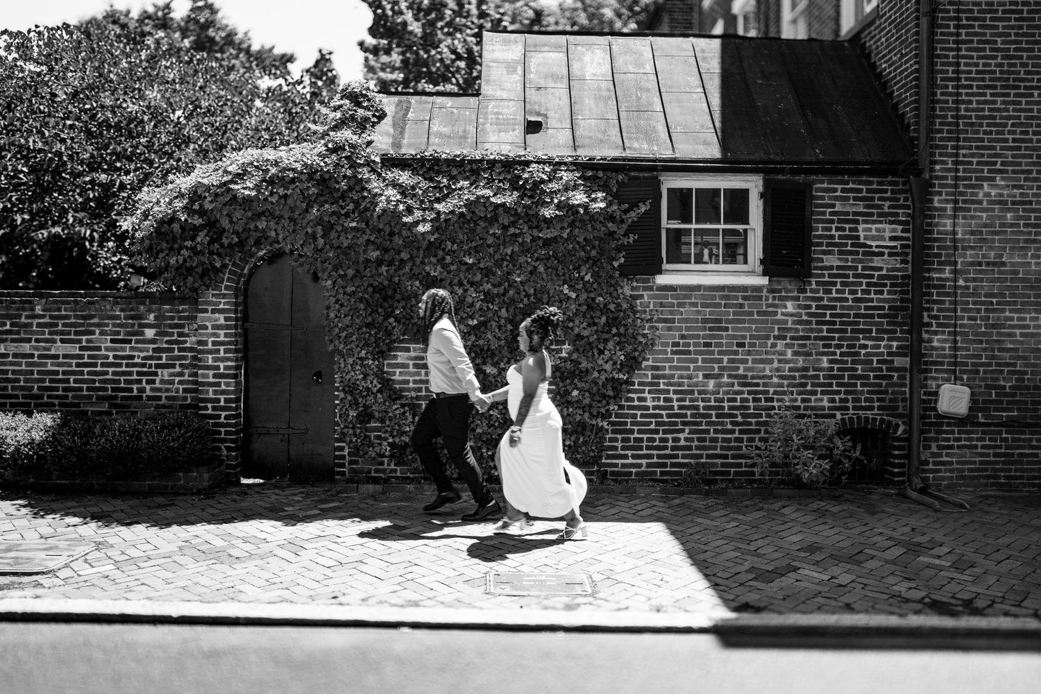 Black and white photo of a bride and groom running along a cobblestone path beside a historic brick building.