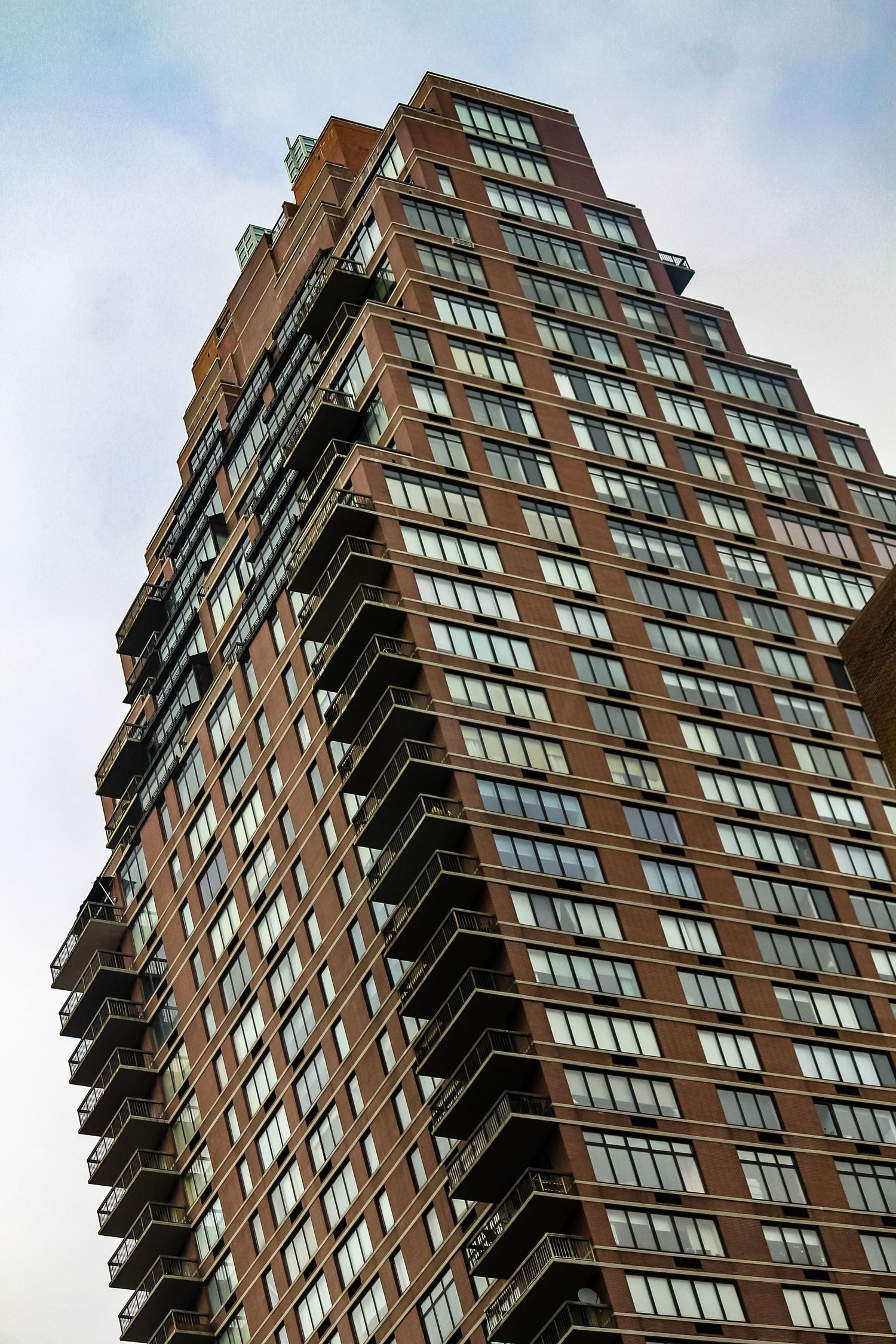 Tall red brick residential skyscraper with rows of windows and balconies against a cloudy sky.