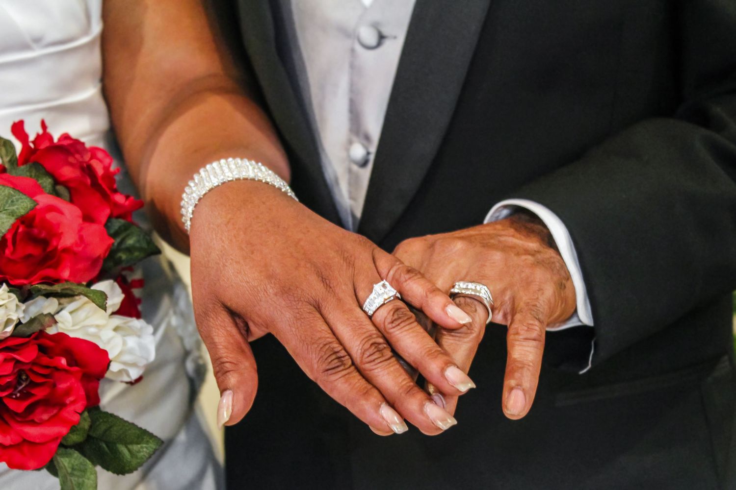 Close-up of newlyweds hands showing wedding rings with red roses bouquet and pearl bracelet.
