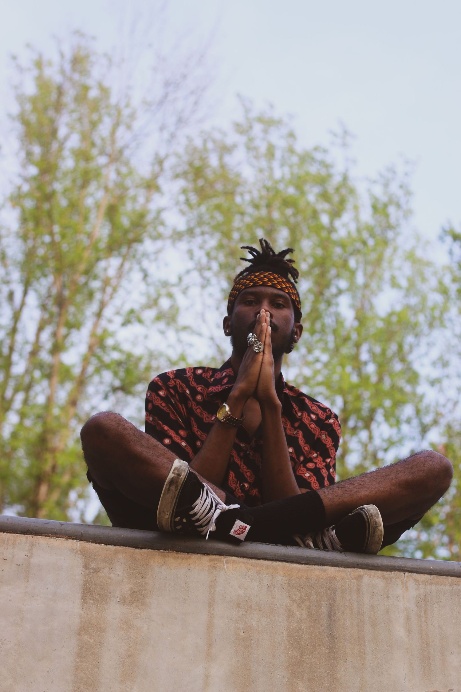 A Black man in a patterned shirt sits cross-legged in a prayer pose on a concrete ledge outdoors.