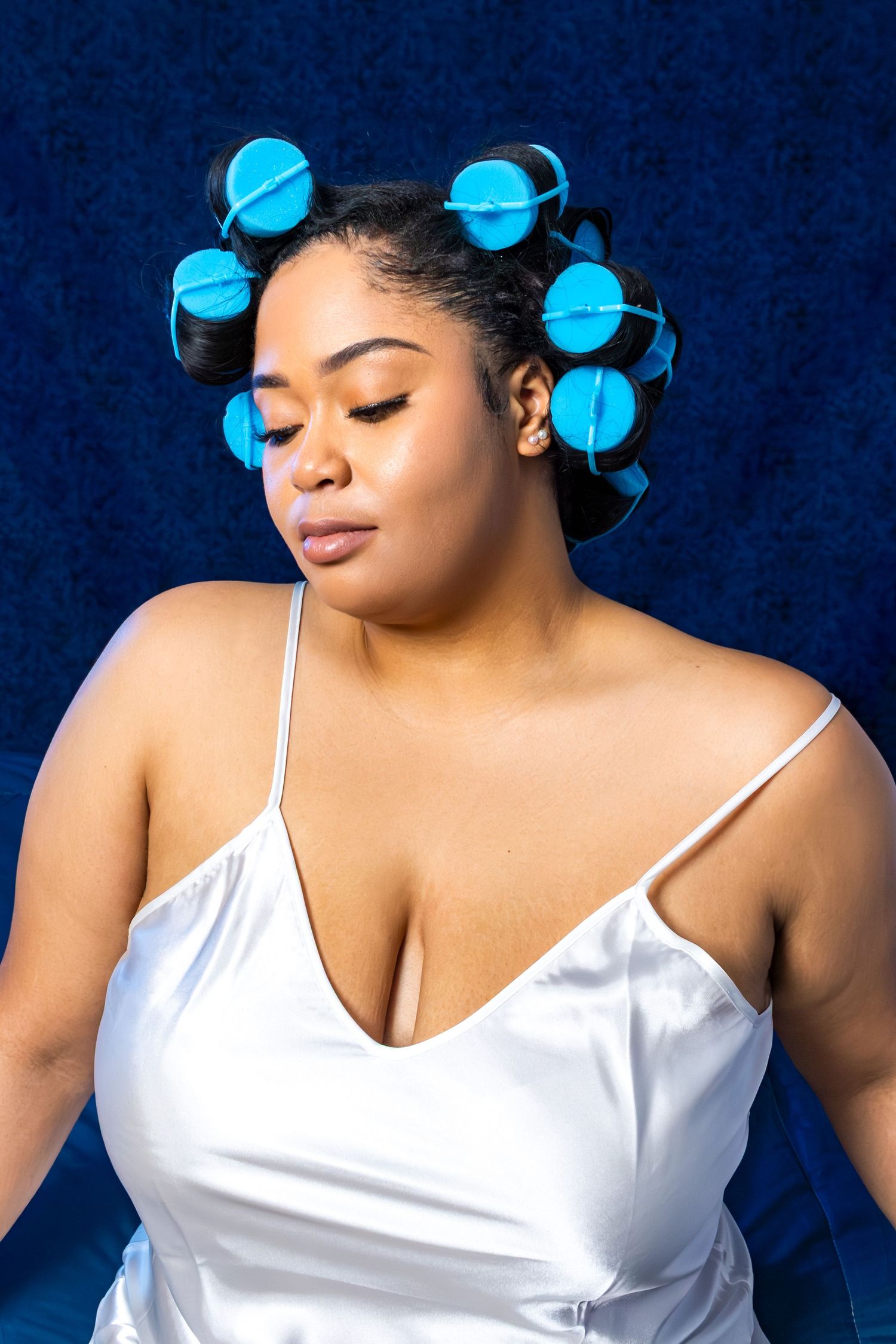 Woman with blue hair rollers wearing a white satin slip dress poses against a dark blue background.