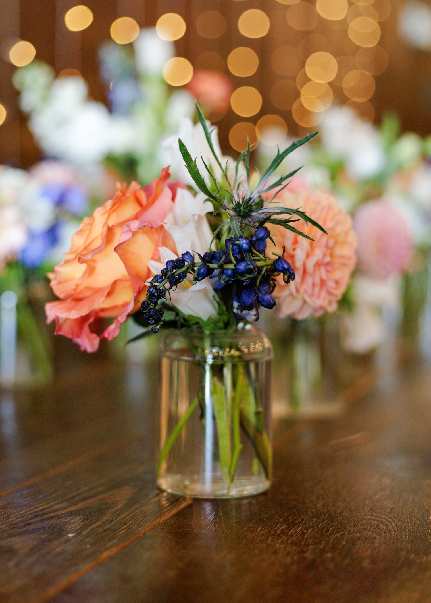 Small glass vases with colorful peach and blue wildflower wedding centerpieces on wooden table.