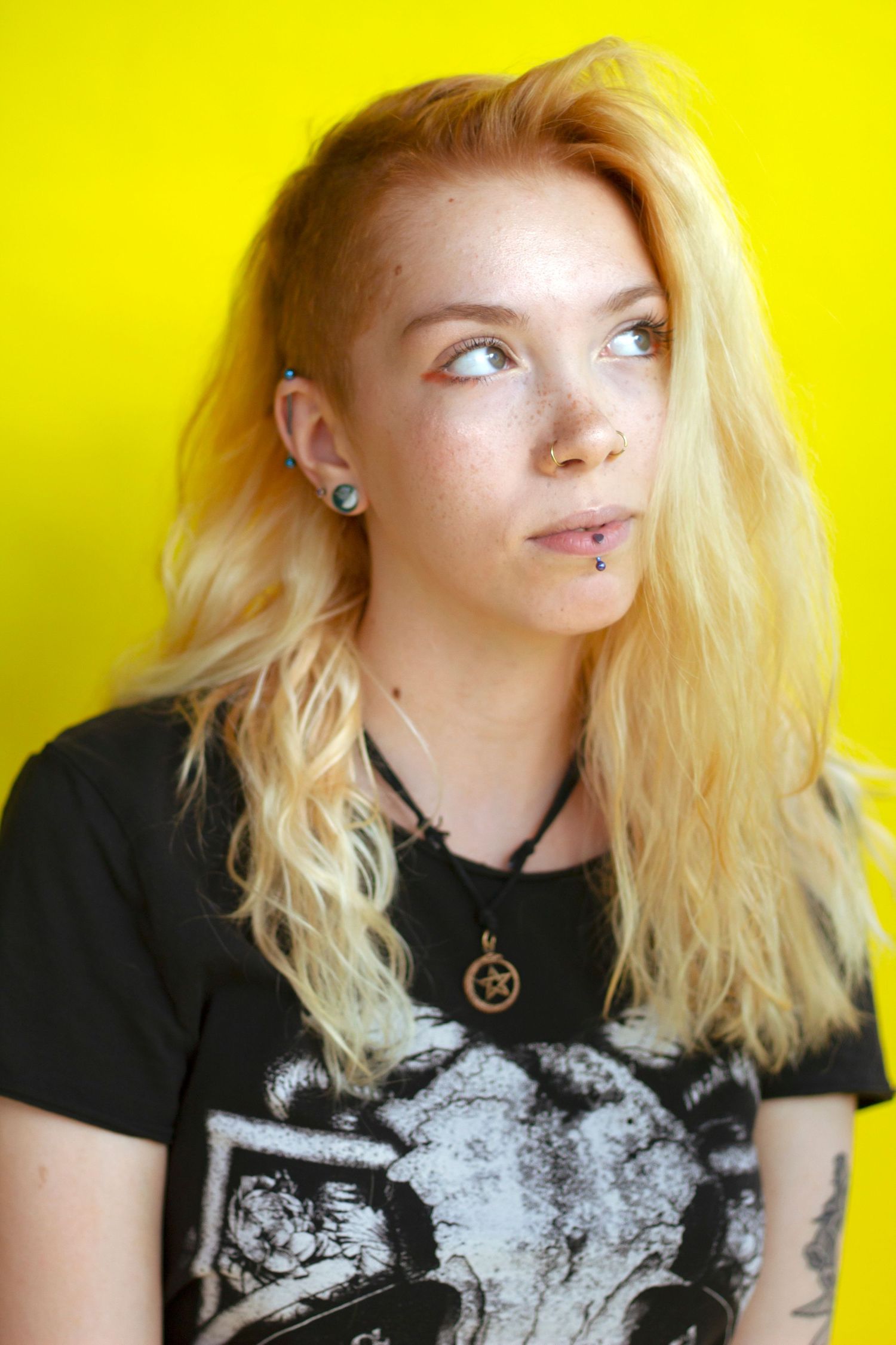Blonde woman with facial piercings and undercut wearing black graphic tee on yellow background.