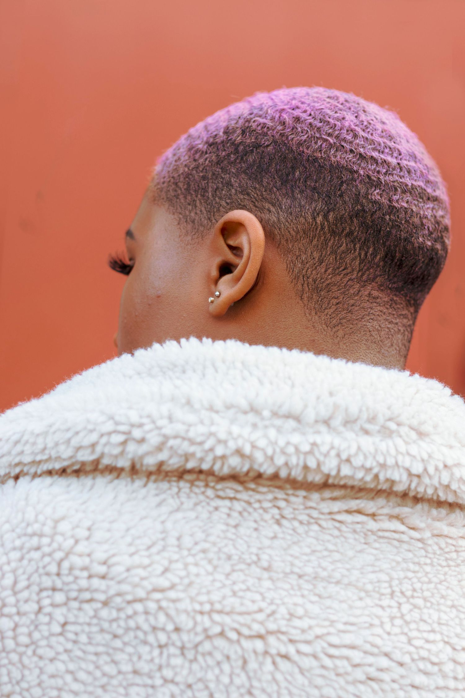 Back view of Black woman with pink buzzcut wearing white sherpa coat against orange-red background.