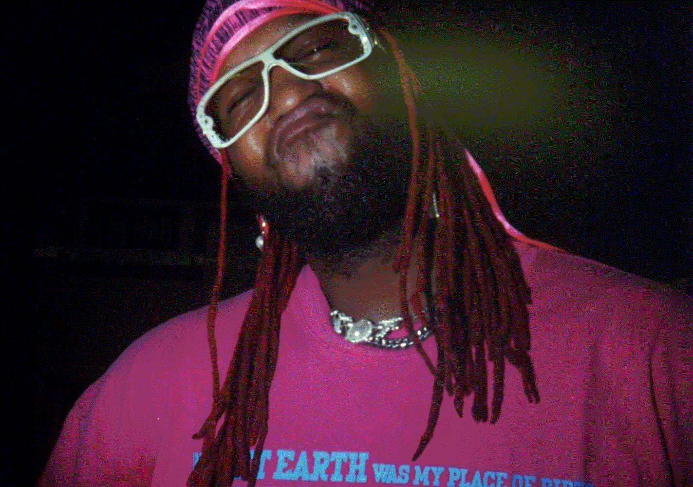 Man with locs wearing pink shirt and white sunglasses against dark background.