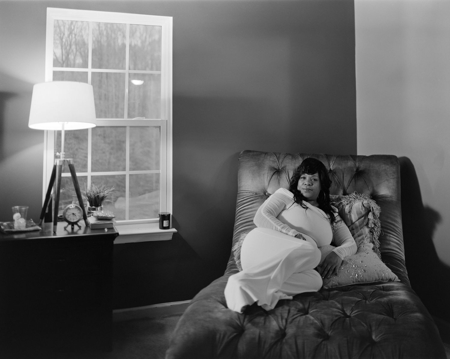 Black and white portrait of pregnant woman in white dress relaxing in armchair by window.