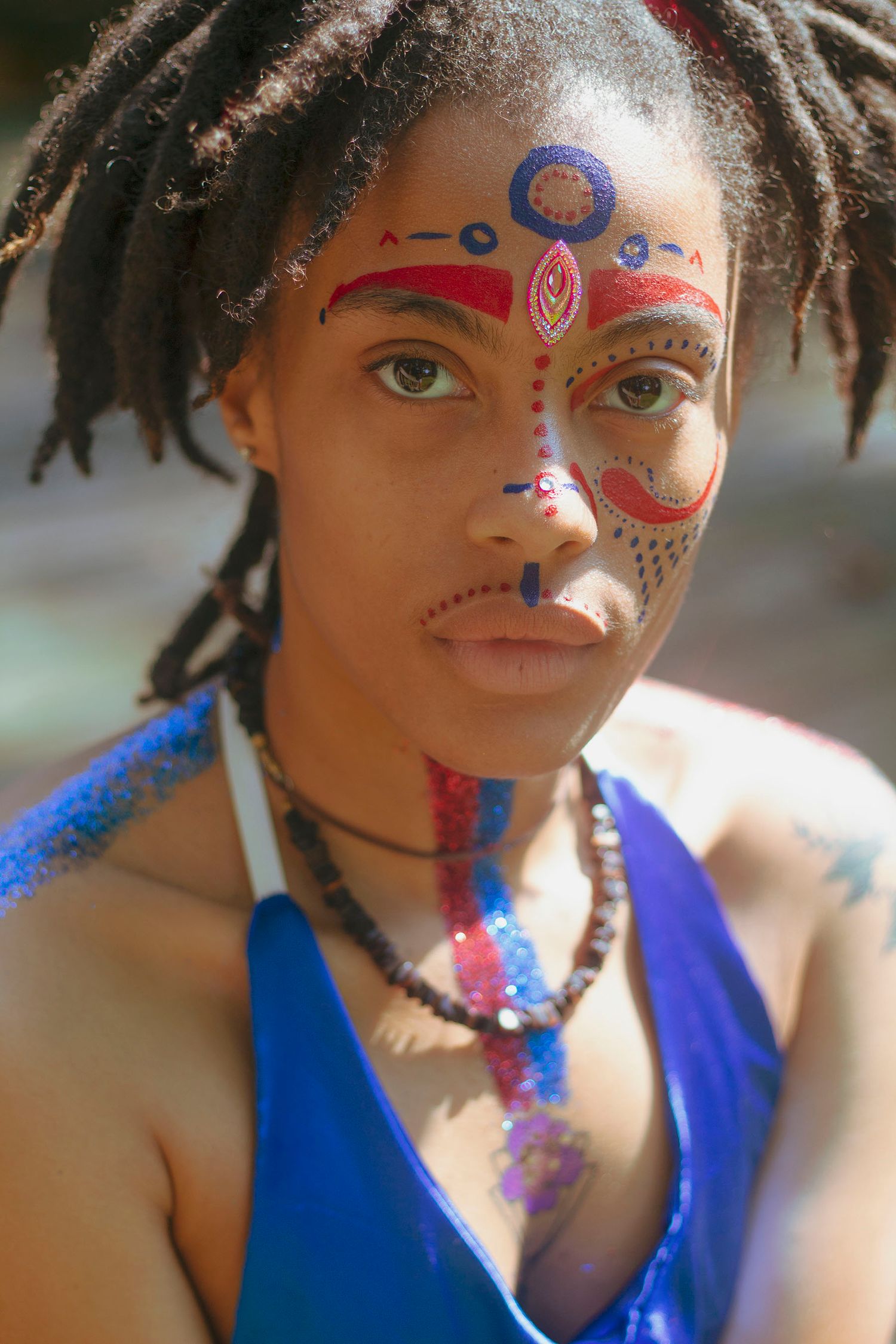 A young Black woman with dreadlocks wears vibrant tribal face paint in red and blue outdoors.