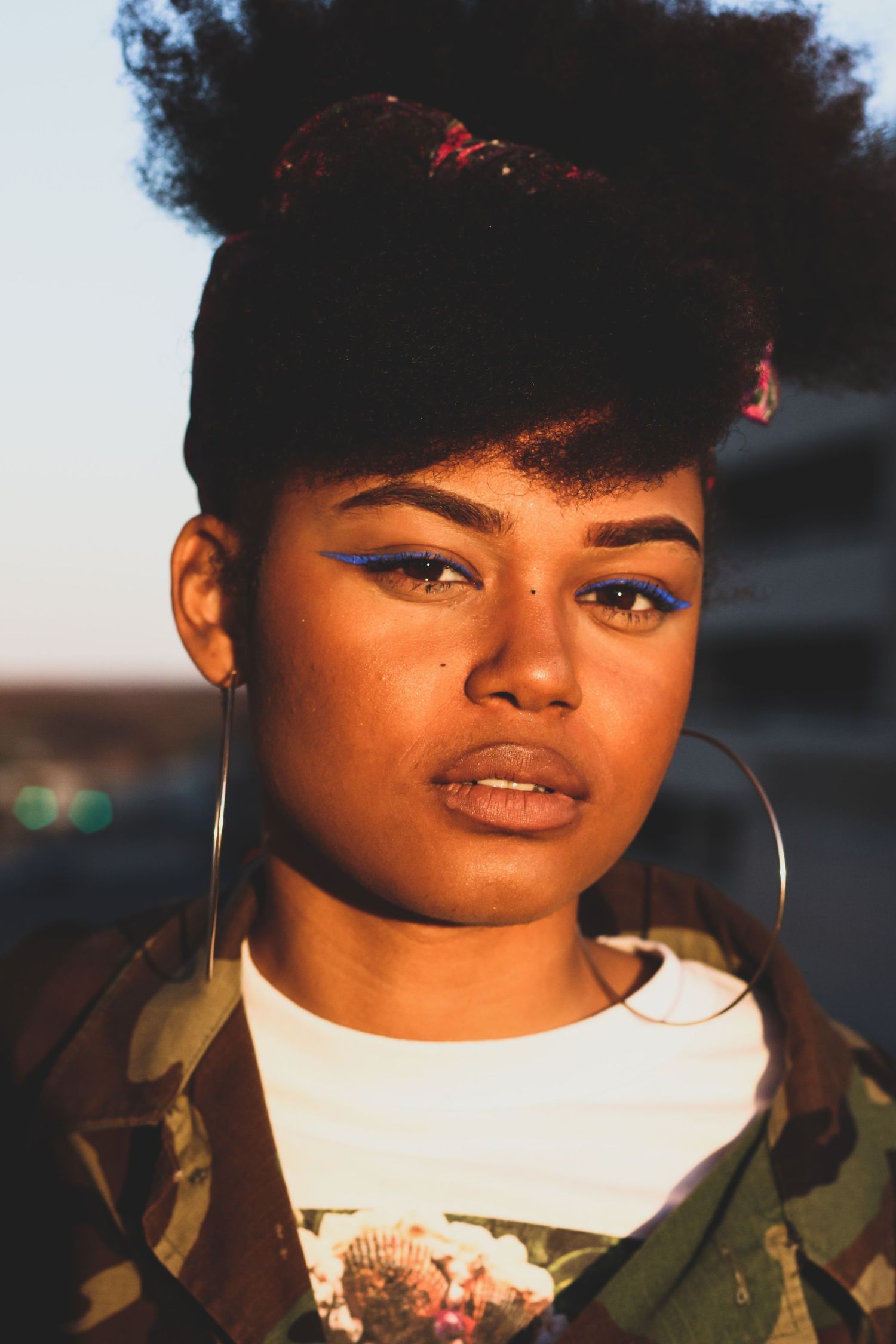 Young Black woman with afro and bold blue eyeliner wearing large hoop earrings outdoors.