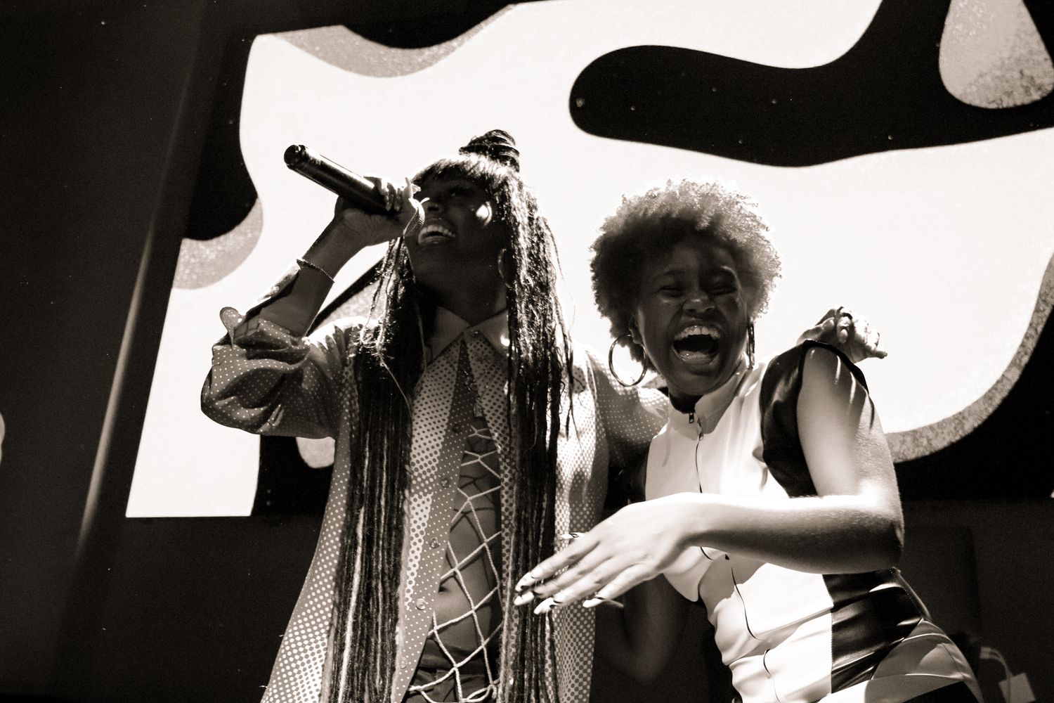 Two female performers sharing a microphone on stage in black and white concert photography.
