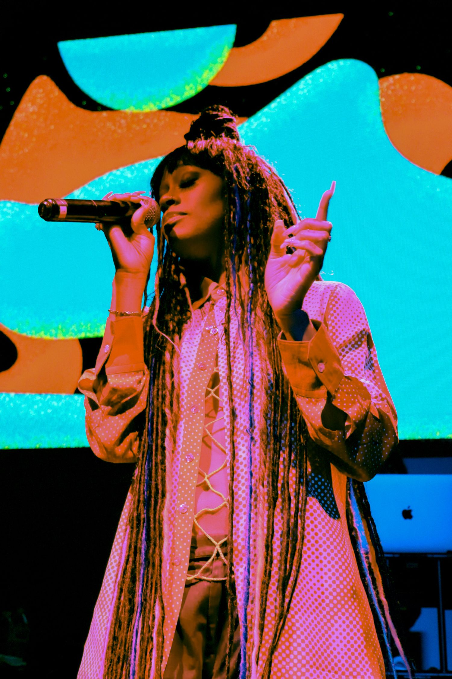Female artist with long braids singing on stage under colorful pink and teal concert lighting.