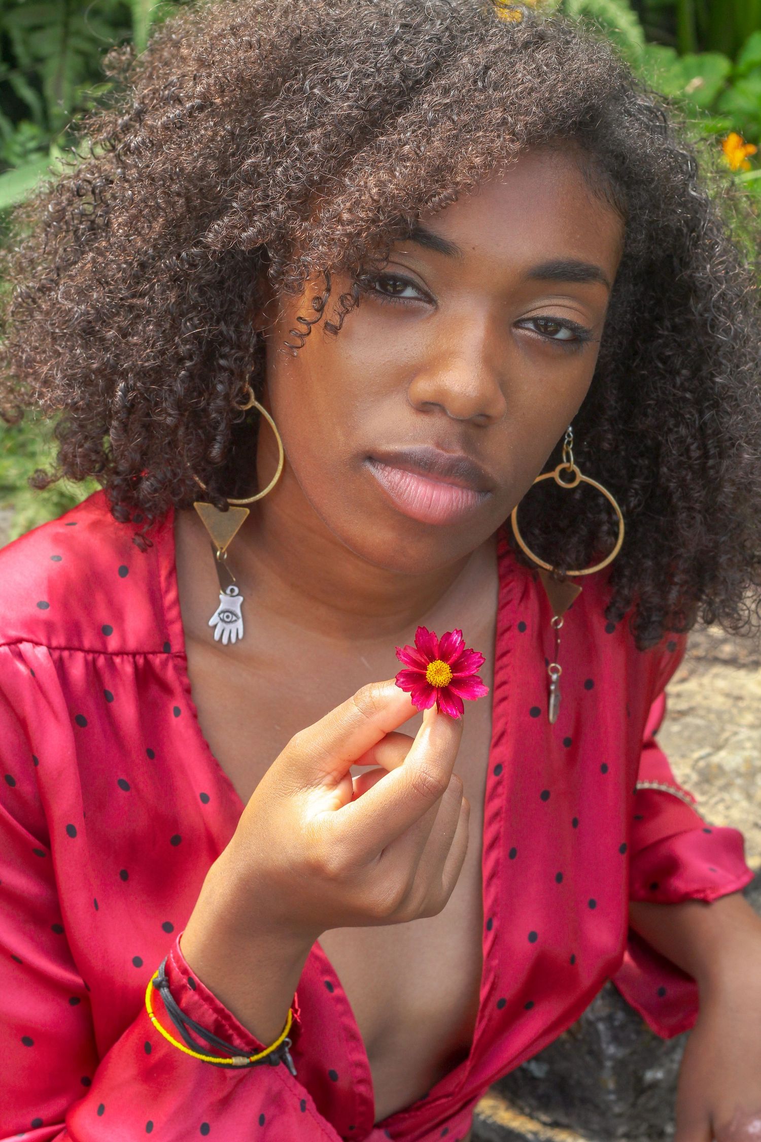 Black woman with curly hair in red dress holding a small pink flower outdoors.