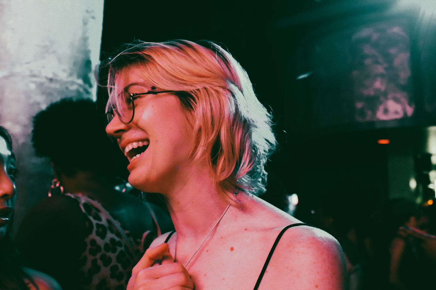 Smiling blonde woman with glasses laughing at a dimly lit social event.