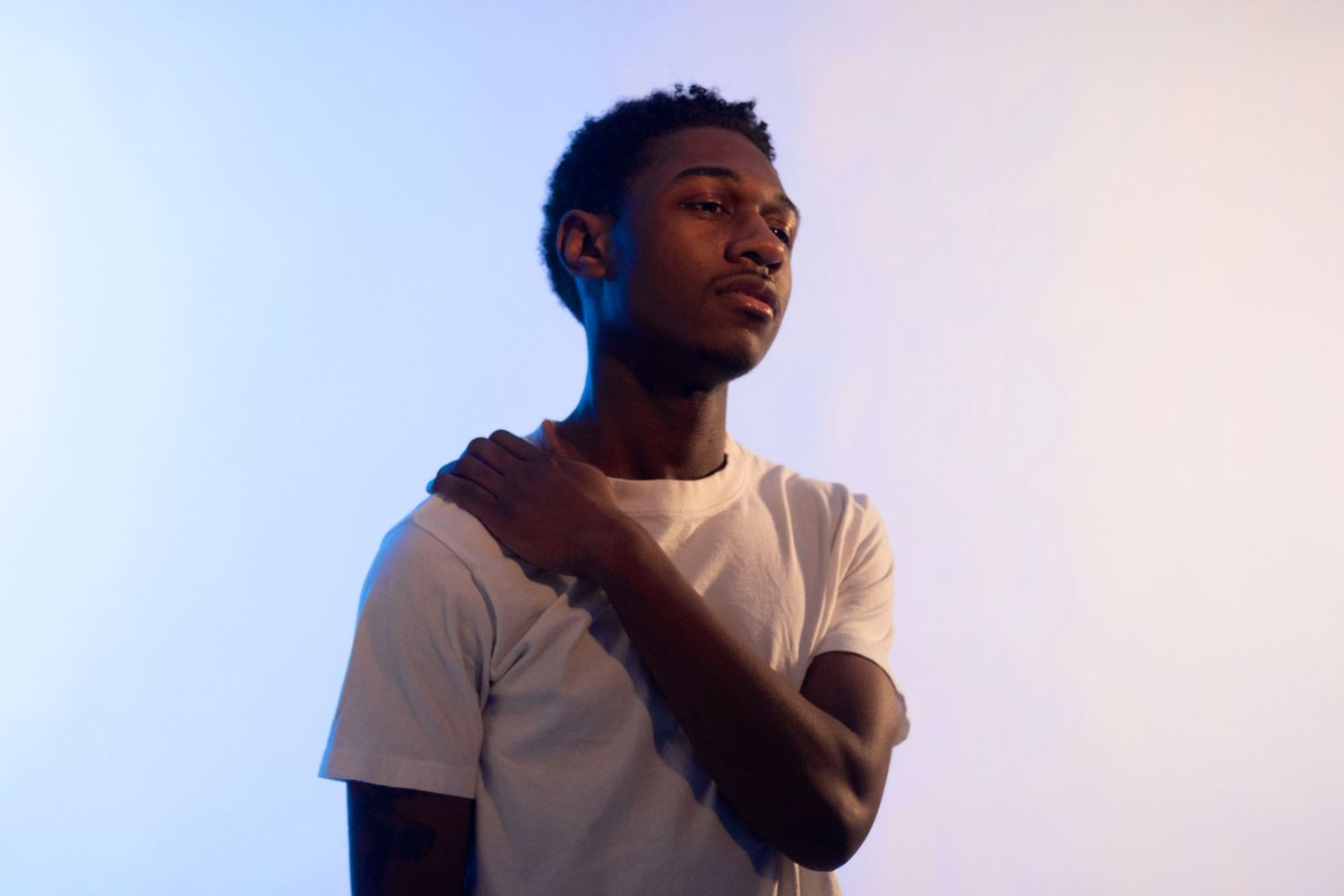 Young Black man in white t-shirt poses against blue-lit studio background with hand on shoulder.