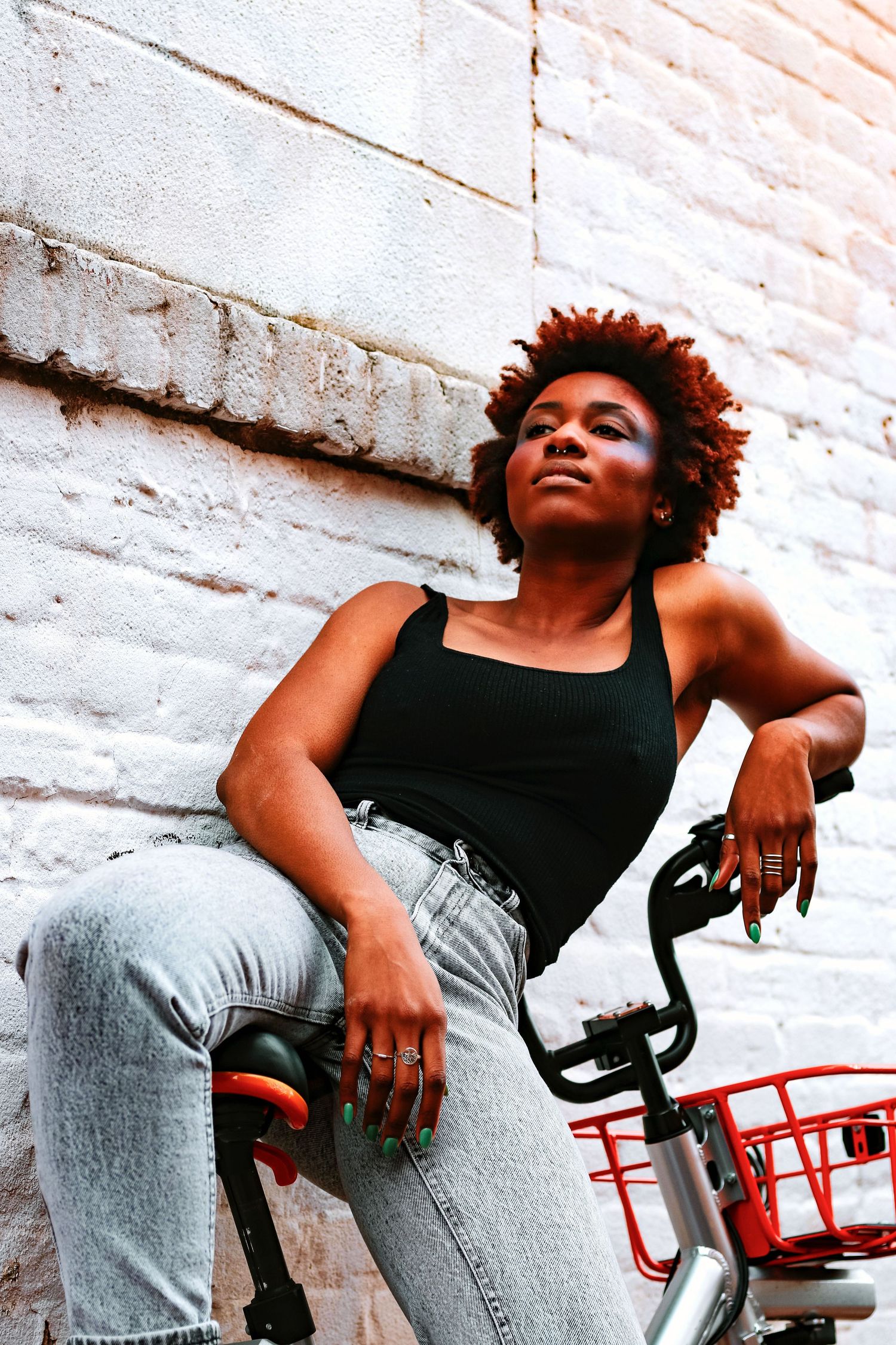A Black woman with auburn natural hair sits on a red bike against a white brick wall in jeans.