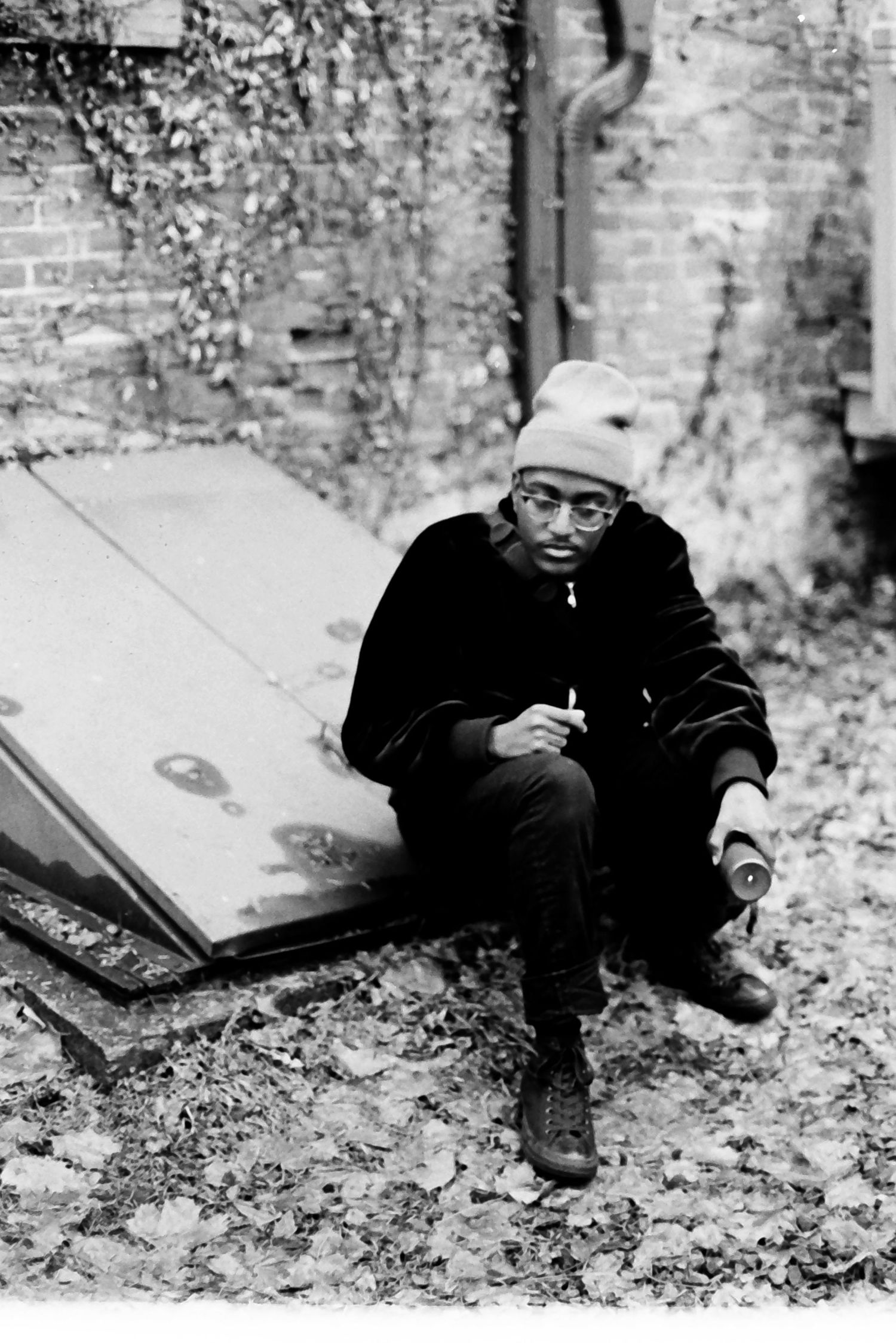 Black and white portrait of person in beanie and glasses crouching outdoors near abandoned wall.